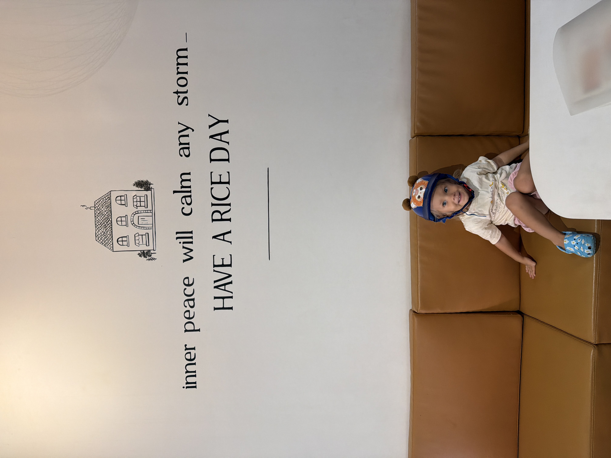 A young girl with blonde hair and blue eyes, wearing a light-colored t-shirt, pink shorts, a blue helmet with cartoon ears, and blue patterned Crocs, sits on a tan leather couch. She is smiling and looking towards the camera. Behind her, on a white wall, is a black drawing of a house and the text "inner peace will calm any storm — HAVE A RICE DAY". The couch is in what appears to be a cafe or restaurant, possibly in Nam Nha Trang Ward, Vietnam, given the context. A white table is partially visible in the foreground.