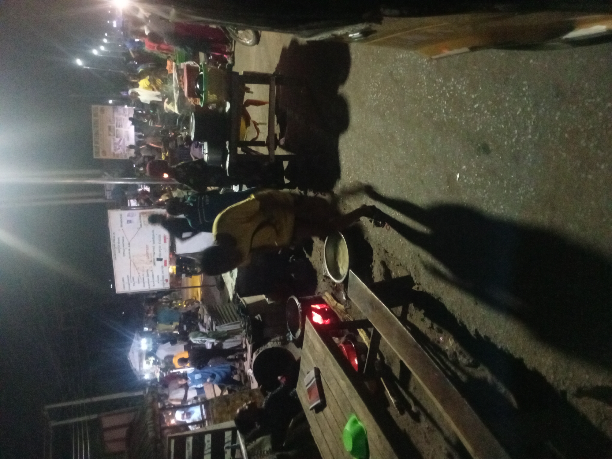 This night-time photograph captures a vibrant street scene in Lokoja, Nigeria, bustling with activity under artificial illumination. Numerous people are gathered, suggesting an evening market or street food gathering.
In the foreground and mid-ground, a central figure wearing a yellow top is actively engaged in cooking or preparing food at a makeshift stall. This stall features a cooking setup with what appears to be hot coals or an open flame, indicated by glowing red embers beneath several metal pots and containers. The cooking station is constructed on a wooden table or platform, placed directly beside a paved road or thoroughfare. Long shadows are cast across the ground due to the strong overhead lights.
The scene is crowded with many individuals, some standing, others seemingly seated or moving through the area. Their silhouettes and forms are illuminated by bright streetlights and stall lights, which create significant glare in various parts of the image.
On the left side of the frame, two prominent signs are visible. The larger, upper sign is white with dark text; although difficult to read entirely, it appears to contain words such as "YOUTH," possibly indicating a "YOUTH PROGRAMME" or "YOUTH EMPOWERMENT" initiative. A smaller sign is situated below it, but its text is illegible. The background shows more people and structures, extending into the distance, further emphasizing the busy, communal atmosphere. The ground is dark, granular pavement, reflecting some of the artificial light. The activity suggests a lively social and commercial environment in an urban setting after dark.