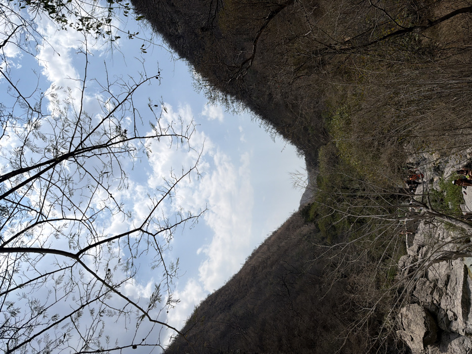 The image depicts a natural valley or canyon landscape in San Miguel Atlapulco, Mexico, under a bright, partly cloudy sky. The perspective is from an elevated position looking down into the valley. Steep, dry, vegetated hillsides frame the view, with the left slope appearing darker in shadow and the right slope illuminated. Dominant vegetation on the hillsides consists of deciduous trees and shrubs with minimal foliage, suggesting a dry season or climate. The foreground and midground feature a rocky terrain composed of large, light-gray boulders and smaller rocks, indicating a streambed or canyon floor.
Bare tree branches, largely devoid of leaves, are silhouetted against the sky in the upper portion of the frame. In the lower-mid section of the image, approximately ten to fifteen individuals are gathered among the rocks. Two individuals are seated on a large rock in the immediate foreground, facing away from the viewer. Further back, a larger group of people is visible standing and sitting on and around the rocks, positioned in what appears to be a natural gathering area. Their attire suggests recreational activity. Lush green foliage is present deeper within the valley, contrasting with the drier vegetation on the slopes. No specific dynamic actions or discernible interactions among the individuals are evident beyond their collective presence in the rocky environment.