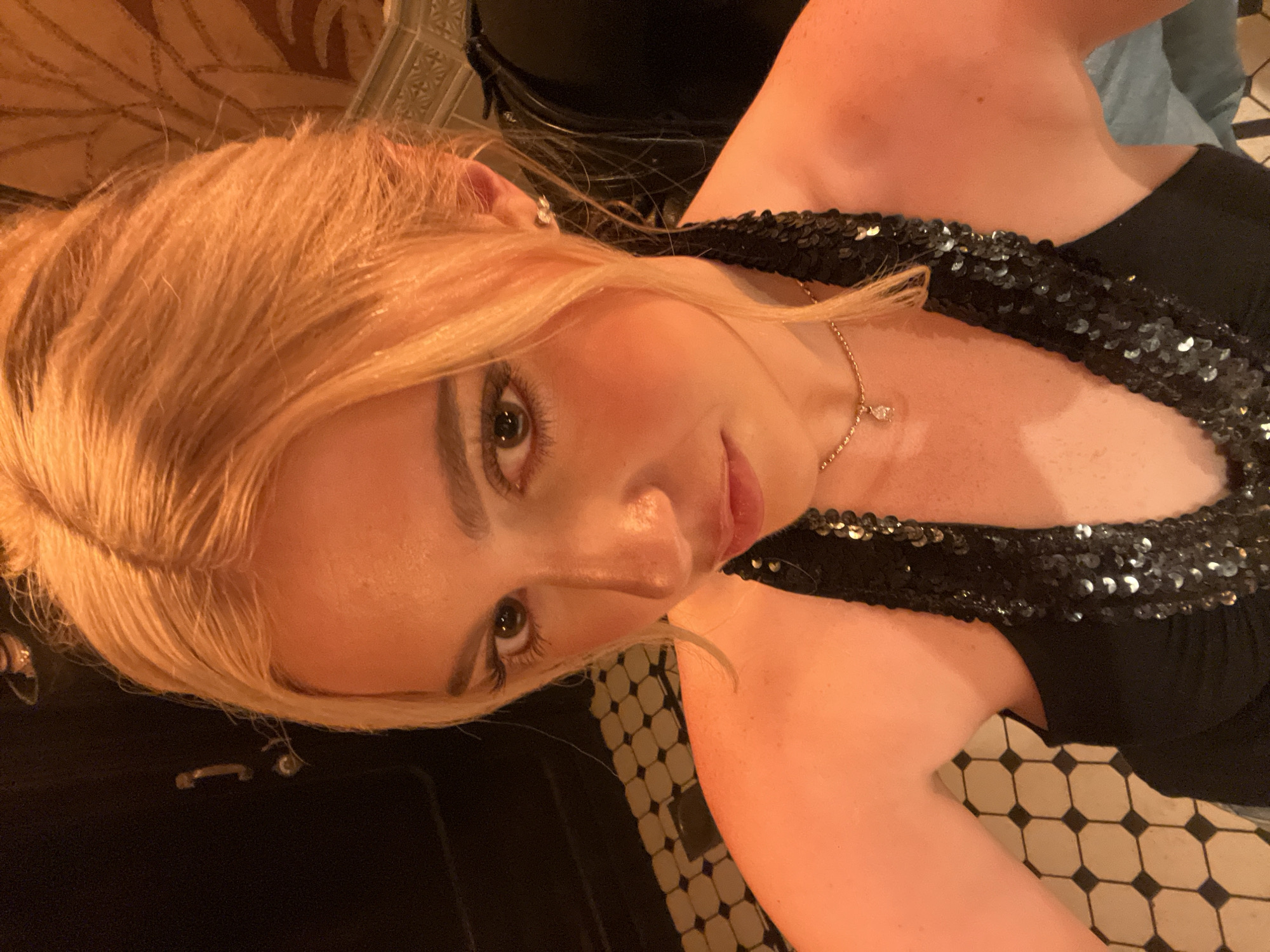 A fair-skinned woman with strawberry-blonde hair gazes directly at the camera, her head tilted slightly. She wears a black dress with a sequined neckline and a delicate gold necklace. The background reveals black-and-white geometric tile flooring and a wall with ornate molding. While the location cannot be confirmed as being in Paris, the architectural details suggest a European setting.