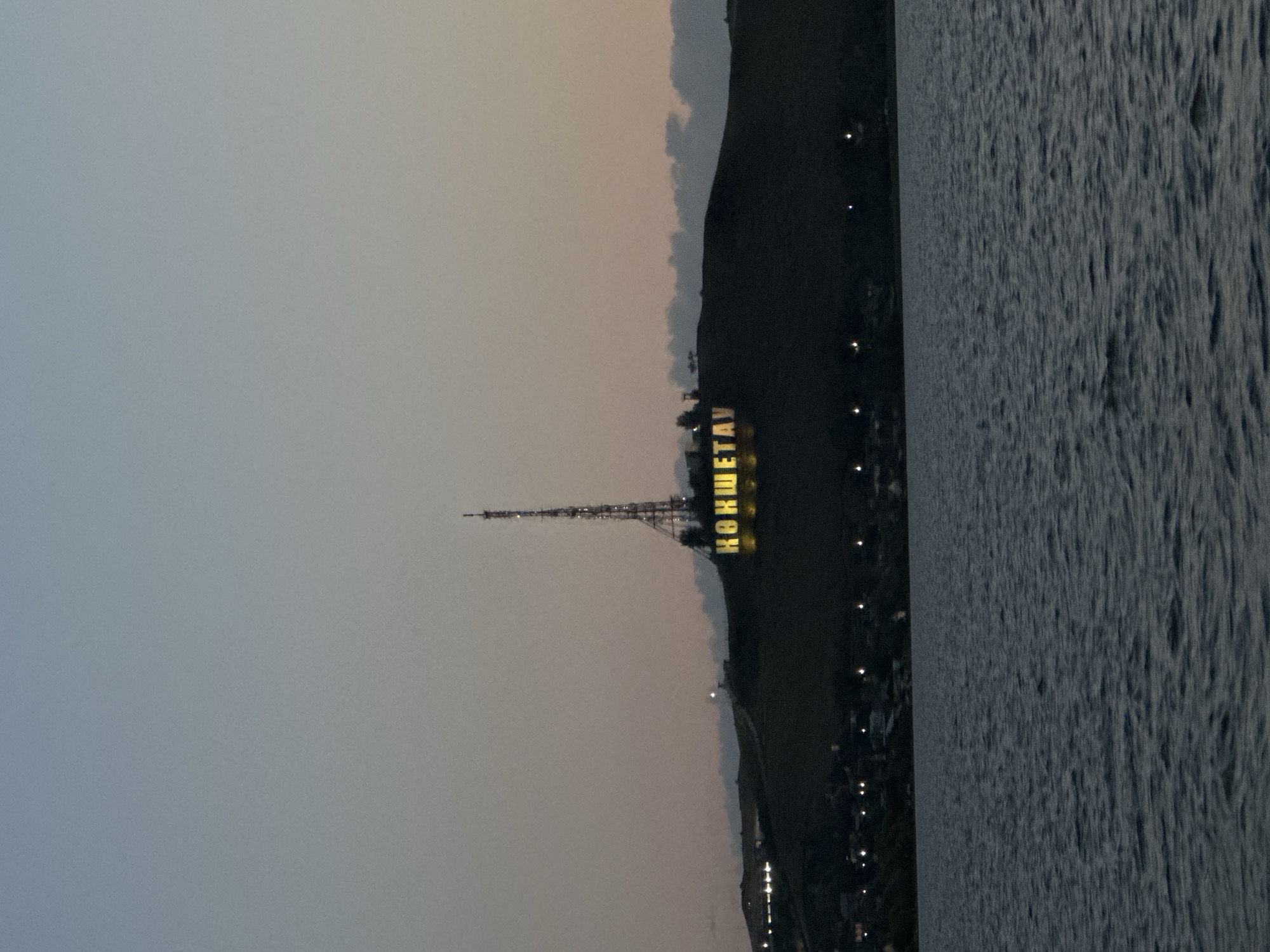The image captures a twilight view of the city of Kokshetau, Kazakhstan. In the foreground, a body of water, likely a lake or reservoir, exhibits gentle ripples. The middle ground features a dark, silhouetted hillside that slopes upwards. Prominently situated on this hill are illuminated letters spelling out "КӨКШЕТАУ" in yellow, likely a landmark or part of the city's name. Towering behind these letters is a tall, lattice-like telecommunications tower, also illuminated. Along the base of the hillside, a faint line of lights indicates the presence of buildings or streetlights within the city. The sky above is a gradient of pale blue and soft orange, suggesting the last moments of daylight or early dawn. No people or animals are visible in the frame.