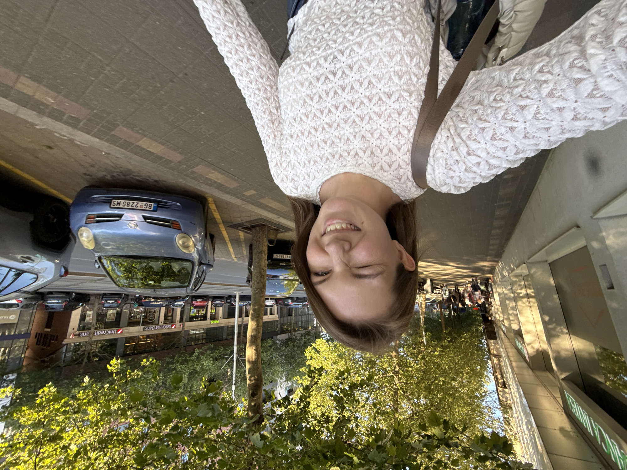A selfie of a woman is oriented upside down. The background reveals an urban scene with a street, parked cars (a blue Nissan with license plate BG 2280-MS), trees, buildings, and other people. The building visible behind the trees is labeled as a Unicredit Bank. This suggests the location is in or near a commercial area. Given the specific location request, it is likely that the location of the photo is near a Unicredit Bank in Vracar Urban Municipality, Serbia.