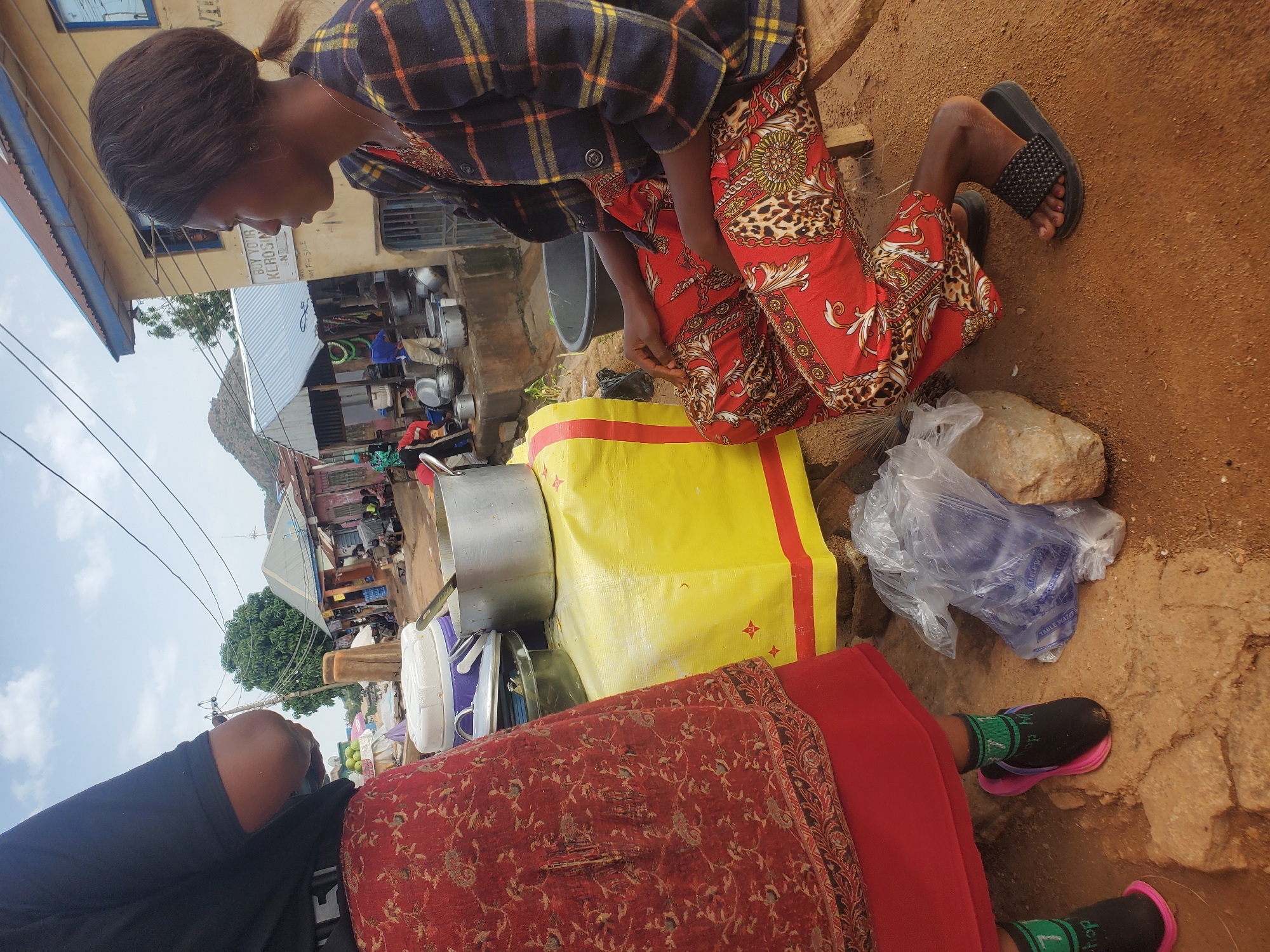 In Gwong, Nigeria, a woman with dark skin and braided hair is seated on a wooden bench. She is wearing a colorful plaid jacket over a red, patterned outfit, and black sandals. To her left, another person, partially visible, is wearing a black shirt and a red, patterned skirt. They are standing near a table covered with a yellow cloth, on which sit several metal pots and containers. In the foreground, a plastic bag filled with water bottles is placed on the dusty ground next to a large rock. The background reveals a marketplace scene with various buildings, stalls, and people going about their daily activities, suggesting a bustling urban environment.