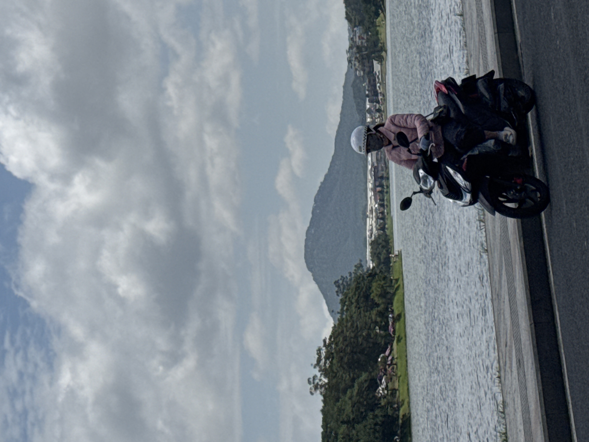 In Da Lat, Vietnam, a person rides a black scooter on a paved path beside a large body of water, likely Xuan Huong Lake. The rider, wearing a pink jacket, a white helmet, and a dark mask, is positioned in the foreground, partially obscuring the view of the lake and the landscape beyond. The lake's surface is rippled, reflecting the overcast sky. In the background, a lush green treeline separates the water from a distant mountain range. The sky is filled with scattered white and gray clouds, suggesting a temperate climate.