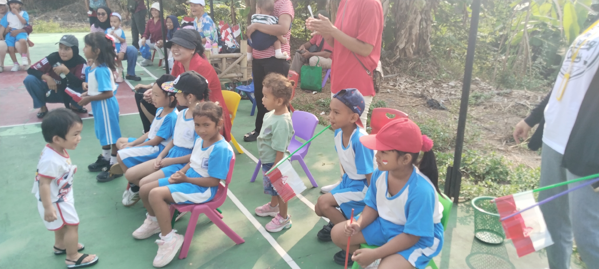 This image depicts an outdoor gathering in Taban, Indonesia, possibly a school event or local celebration. The scene features a diverse group of people, primarily children and adults, engaged in various activities. Several children are seated on colorful plastic chairs, wearing blue and white uniforms, suggesting a school setting. Some are holding small flags, red and white in color, possibly indicative of a national celebration or competition.
One young boy in a white t-shirt and shorts stands in the foreground, facing left. Adults are visible in the background, some standing and observing, while others are seated or interacting with the children. The environment appears to be a paved area, possibly a sports court or playground, with a grassy and wooded area bordering it. Sunlight filters through the trees, creating a bright and warm atmosphere.
Notable objects include the colorful plastic chairs, the small flags held by the children, and a green mesh waste bin. The presence of flags suggests a patriotic or festive occasion. The overall mood appears to be one of communal engagement and activity.