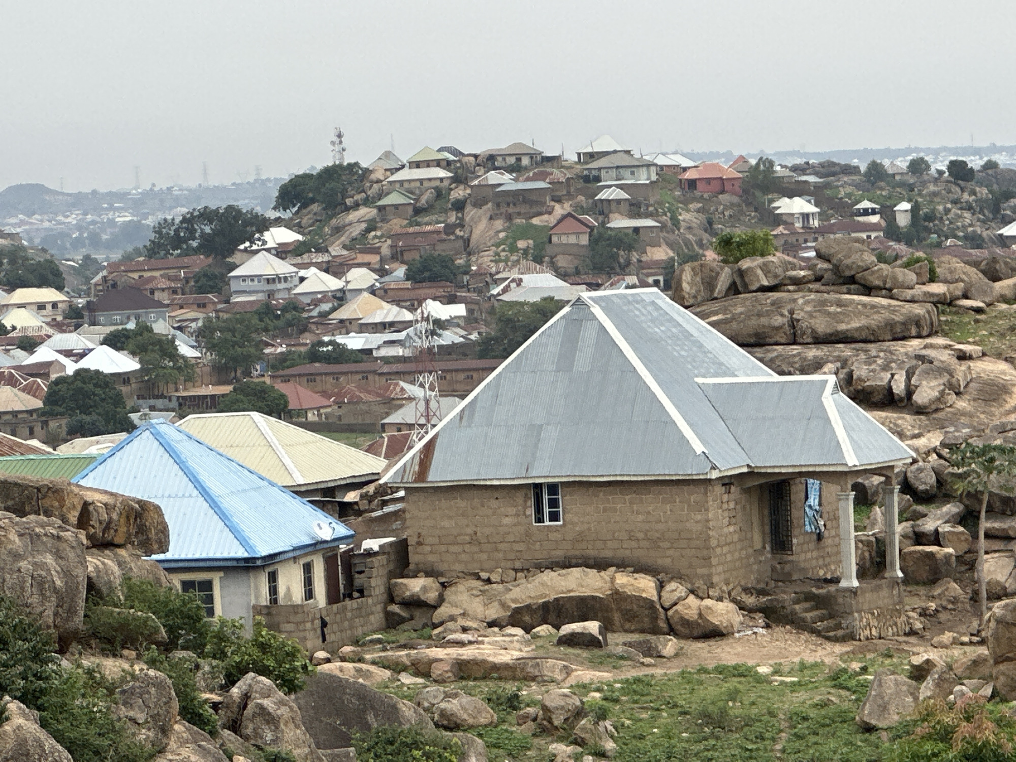 The image shows a settlement in Jos, Nigeria, characterized by a mix of residential buildings nestled among rocky hills. Foreground elements include two houses with corrugated metal roofs, one painted blue and the other unpainted, surrounded by large boulders and sparse vegetation. The houses appear to be constructed from brick or concrete blocks. In the background, a dense cluster of houses with various roof colors and designs is visible, sprawling across the hillside. A communications tower rises amidst the structures. The sky is overcast, casting a muted light over the scene. The environment is rugged, with rocks and vegetation dominating the landscape around the buildings. No people or animals are immediately visible in the image.