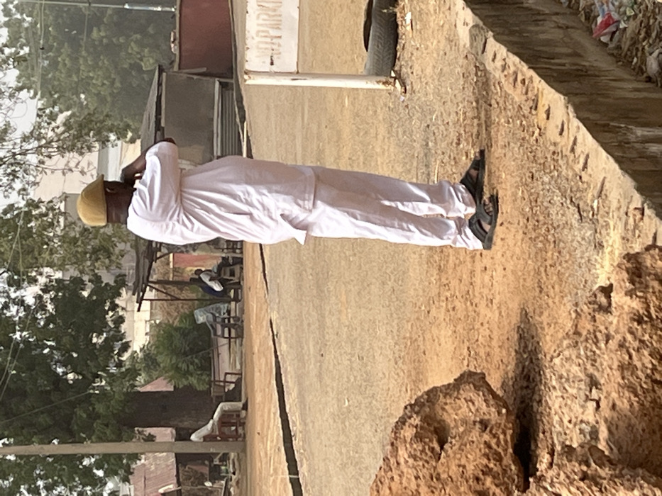 The image depicts an outdoor street scene in Kanti, Nigeria, under clear daylight conditions. In the mid-foreground, a man stands in profile, facing right, wearing a light-colored long-sleeved tunic over matching trousers and black open-toed sandals. He is also wearing a light brown flat cap, and his hands are raised towards his face. To his immediate right, a white sign post is visible, with "NO PARKING" text partially legible. An old tire rests on the dirt ground near the sign.
The ground consists of reddish-brown dirt, with a paved or compacted dirt path leading into the mid-distance. A concrete curb runs along the lower right edge, with some refuse visible beneath it. In the foreground to the left, a large, irregularly shaped brown rock or mound of earth is present.
In the background, beyond the path, several individuals and structures are visible. On the far left, a person sits on a white chair under a large tree with green foliage, positioned next to a utility pole. Further to the right, an open-sided shelter with a dark roof and support poles houses two or three indistinct figures. Other small buildings or shacks with corrugated metal roofs are scattered throughout the background, amidst additional green trees and foliage. The overall lighting suggests a bright, sunny day.