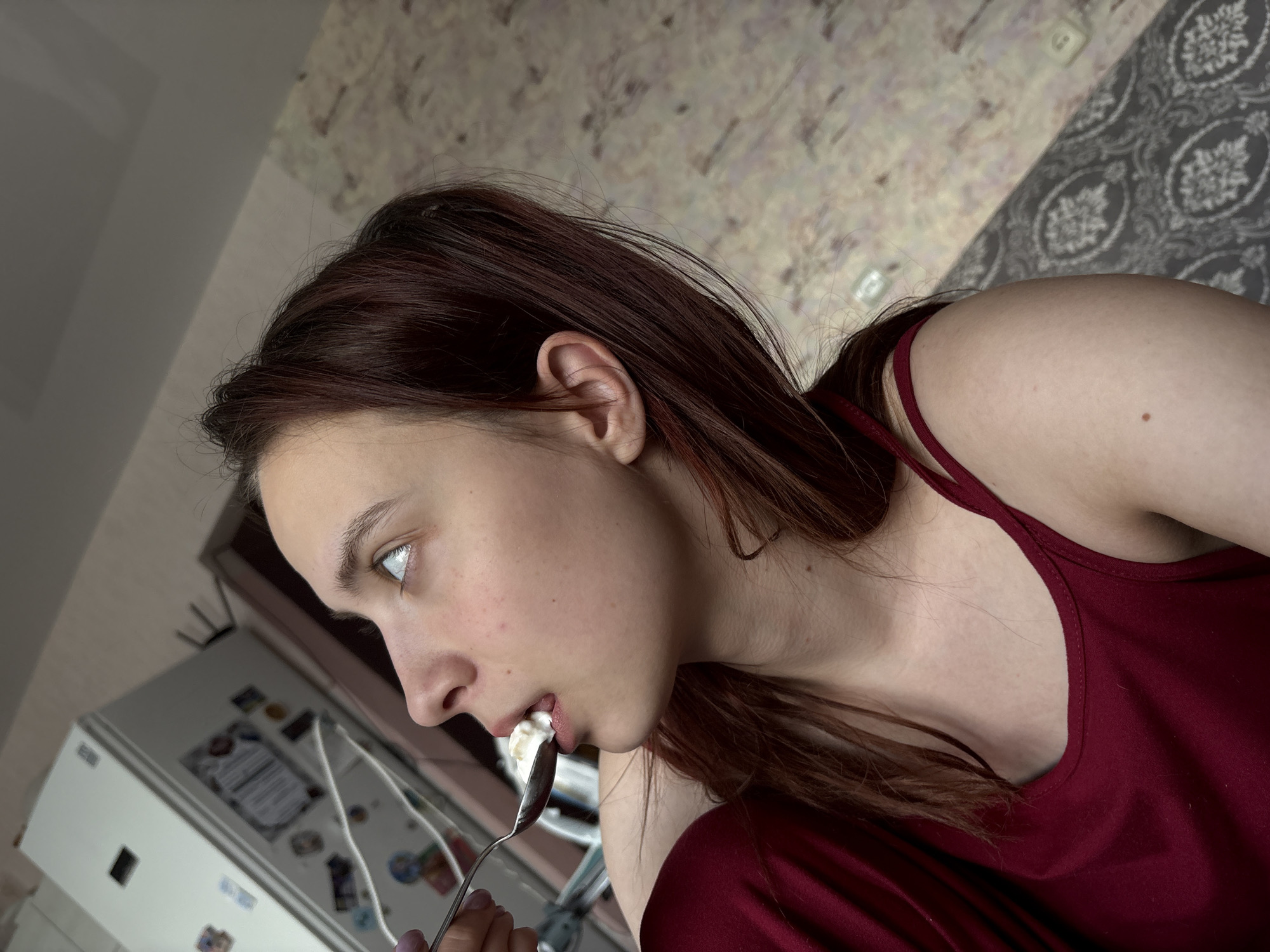 A young woman with dark brown hair, wearing a dark red camisole, is captured in an angled, close-up shot. Her left profile is visible as she eats a white, creamy substance from a silver spoon. Her mouth is slightly open, with the food resting on her lower lip. Her gaze is directed upwards and to the left, indicating focus on the activity.
The setting appears to be indoors, likely a kitchen or dining area within a home. In the background, a white refrigerator, adorned with various magnets and possibly papers, is partially visible and out of focus. Behind her head and shoulder, a wall features patterned wallpaper with a grey background and a lighter, ornate damask-like design. A plain lighter-colored ceiling or upper wall section is also visible.
The lighting suggests daytime, with soft illumination across the scene. The woman's expression is neutral and concentrated on eating. No discernible text is visible on the refrigerator or elsewhere. The image depicts a casual, everyday moment, potentially in a residence in Прикамский, Russia.