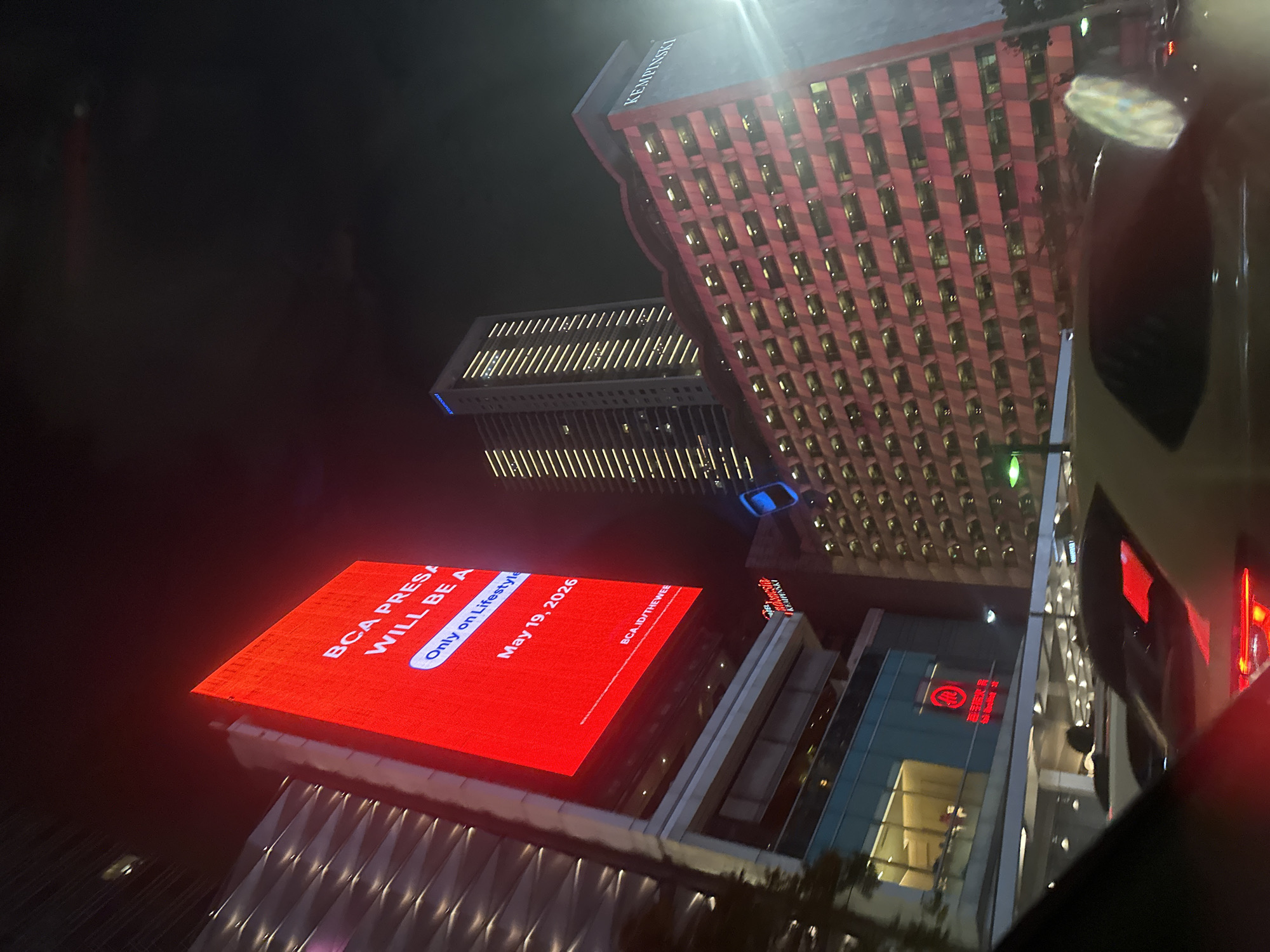 This night shot looks up at buildings in Central Jakarta, Indonesia. The Hotel Indonesia Kempinski stands out as a high-rise with a vertically striped pink and red facade. Nearby, another building glows with rows of yellow lights. A flat rooftop is covered in a bright red LED display with white text: "BCA PRESA WILL BE A", "Only on Lifestyle", "May 19, 2026", and "BCA.ID/THEWEE". Below this is a modern building with glass and lights, including a red circular logo. The viewpoint is from a vehicle.