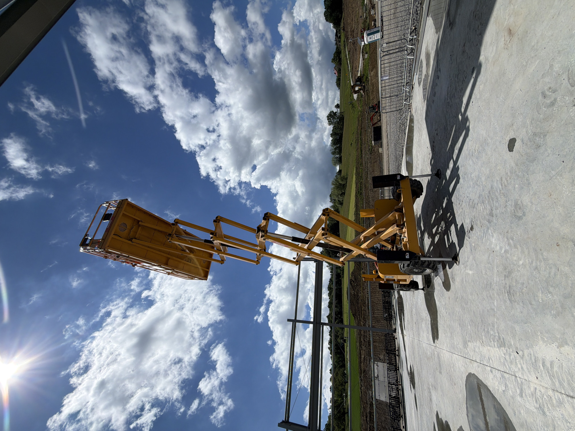 The image shows a bright yellow scissor lift, fully extended, positioned on a concrete surface at a construction site. The lift is in the foreground, angled towards the sky, with its platform at the top. In the background, there is a clear blue sky with scattered white clouds, and the sun is shining brightly, creating a lens flare effect in the upper left corner. Beyond the concrete area, there is a grassy hill with trees and some construction materials, including what appears to be metal beams and a portable toilet. The ground is a mixture of concrete, gravel, and some puddles of water. The overall impression is of an active construction site on a sunny day. The location is identified as Janowice, Poland.