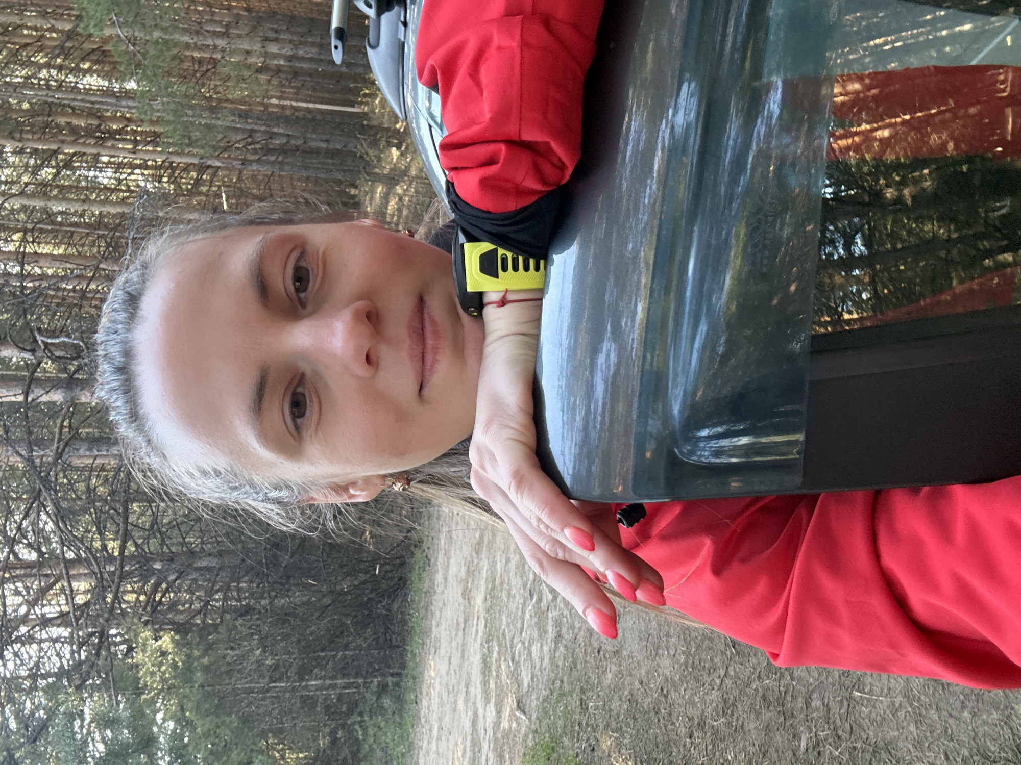 A woman is pictured outdoors against a background of trees, possibly a forest or park. She is wearing a red jacket. Her left hand is resting on a reflective surface, likely a car window. She has fair skin and light-colored hair. Her fingernails are painted pink. A yellow object is visible near her wrist. Based on the visual information, I cannot confirm the location as Chaikovsky, Russia, nor provide any forensic or interaction details.