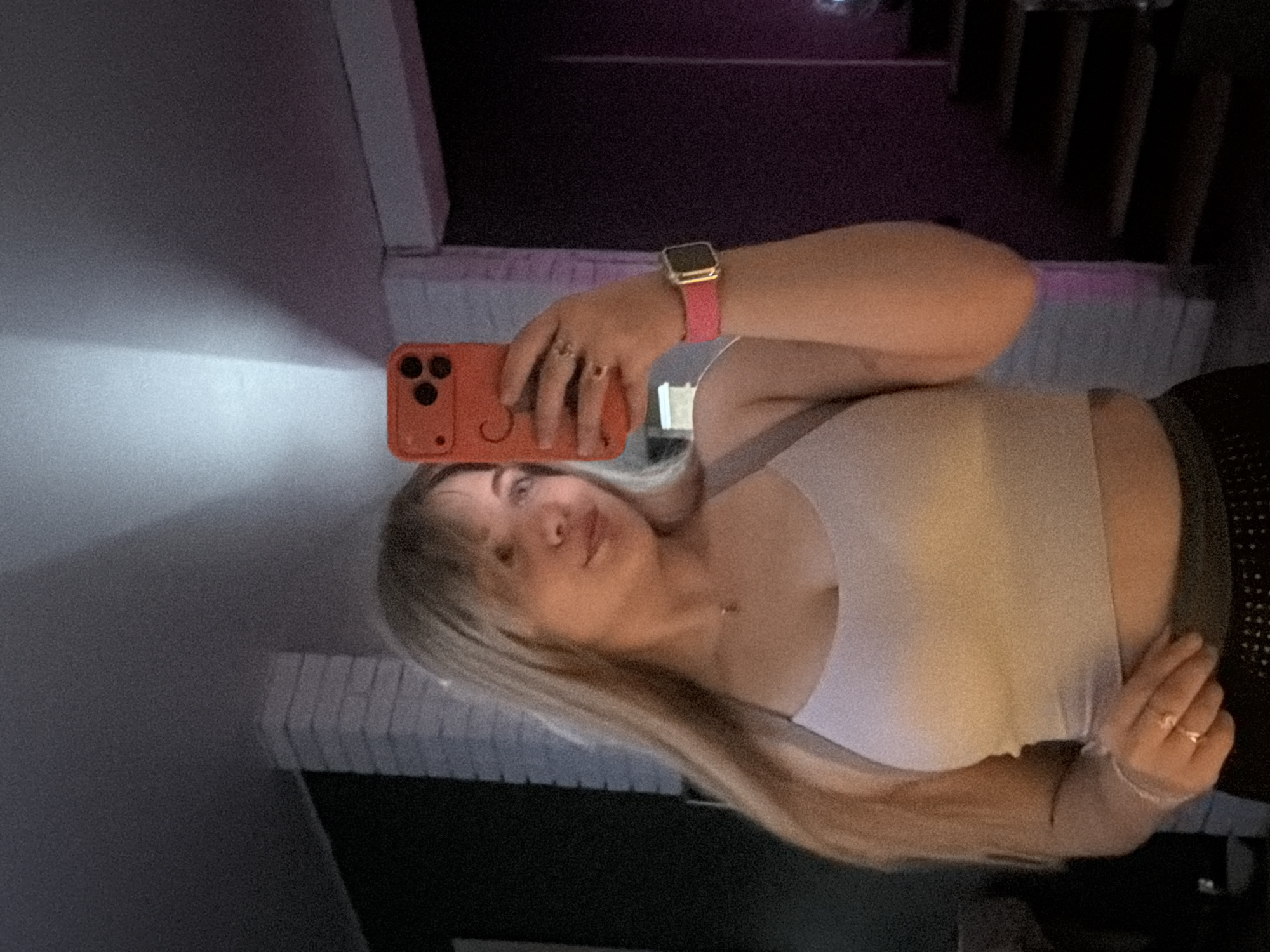 A woman is holding an orange phone while taking a selfie indoors. She is wearing a white top and a pink Apple watch. The location is the city of Большая Шильна, Russia.
