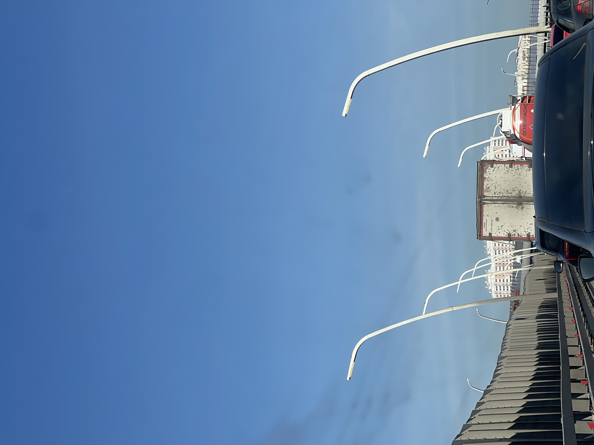 The image shows an elevated roadway lined with modern streetlights in Moscow, Russia. The view appears to be from a vehicle traveling on the road, with other vehicles including a tank truck also present. Buildings are visible in the distance. The sky is clear and blue.