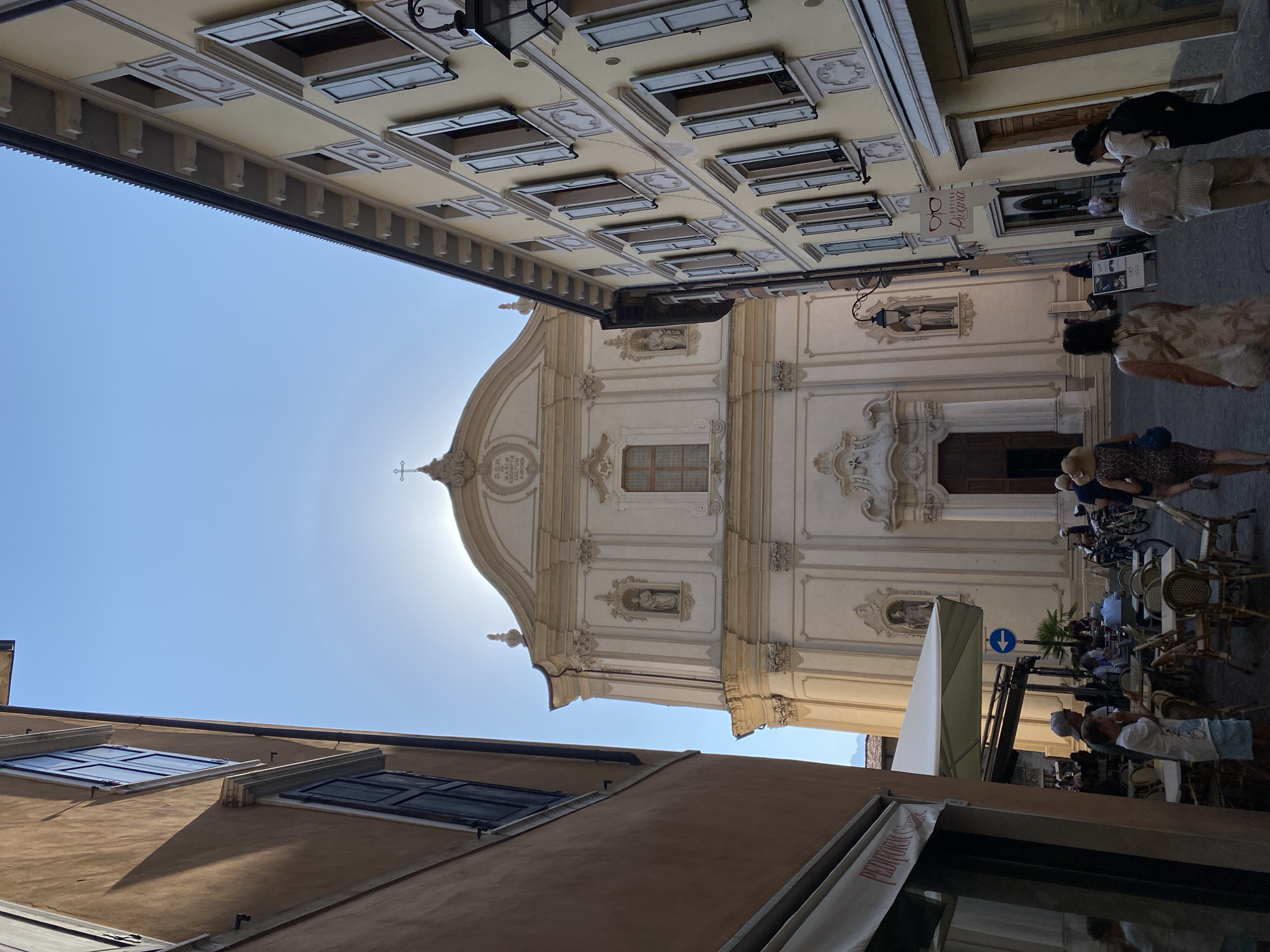 The image shows a view looking up a narrow street towards a church in Riva del Garda, Italy. The church facade is white and ornate, with a cross at the top. The sun is bright and positioned behind the church, creating a halo effect. On either side of the street are buildings with pale yellow and tan facades, featuring multiple windows with shutters. Some people are visible in the street, walking and sitting at outdoor tables of a cafe. The ground is paved with cobblestones. A street lamp is attached to the building on the right. A sign for "Ottica Pigoni" is also visible.