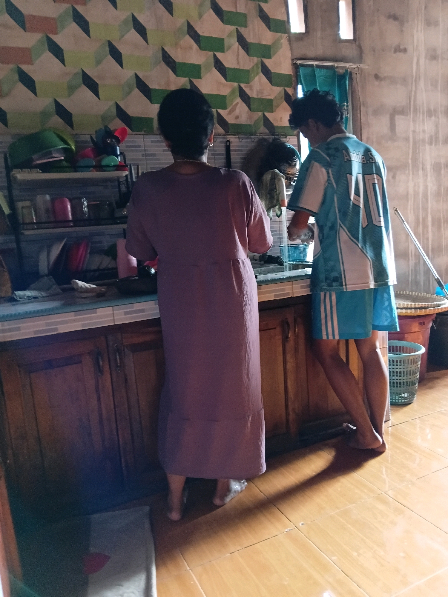 In a modest kitchen located in Margo Rukun, Indonesia, two individuals are engaged in a domestic activity. A woman, viewed from behind, stands to the left, wearing a long, dusty rose-colored dress that reaches her ankles. Her dark hair is styled neatly, and she is adorned with a thin necklace. To her right, a younger person, also seen from the back, is dressed in a blue and white jersey with the number "40" on the back, and matching blue shorts. This individual is leaning over a kitchen counter, appearing to be washing dishes or performing a similar task at the sink. The kitchen environment is characterized by wooden cabinets below the counter, a tiled backsplash, and a patterned wallpaper on the wall behind them. Various kitchen utensils and containers are visible on shelves above the counter. The floor is made of light-colored tiles, and the scene is illuminated by natural light filtering in from windows. To the right of the scene, a large woven basket and a smaller green plastic basket are visible, suggesting a casual and practical living space.