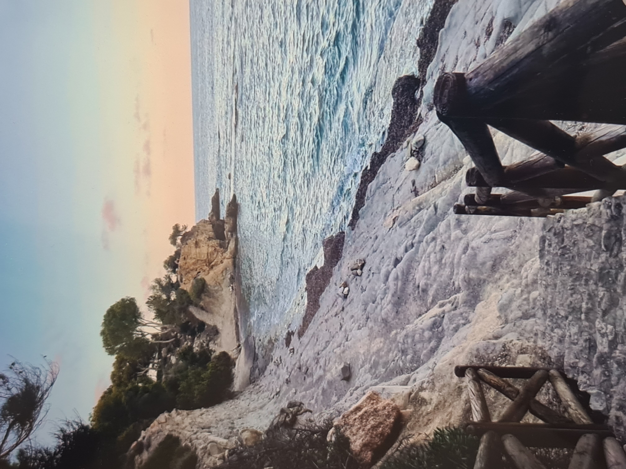 The image displays a picturesque coastal landscape, likely captured during sunrise or sunset, evidenced by the sky's soft gradient from light blue to warm peach and orange hues. The perspective is from an elevated rocky path or viewpoint looking along the coastline.
In the foreground on the right, a rustic wooden railing, constructed from interconnected logs, frames the view, suggesting a marked path or overlook. The ground immediately around it is composed of light-colored, textured rock formations.
The midground features a calm sea with gentle ripples, appearing a clear turquoise-blue. It meets a rugged, rocky shoreline. Further along the coast to the left, a headland covered in dense green trees and bushes, including some distinctive wind-swept trees, extends into the sea. At the base of these cliffs, a small, secluded sandy beach or cove is visible.
No people or visible text are present in the image. The scene conveys a sense of tranquility and natural beauty under clear, pleasant weather conditions.