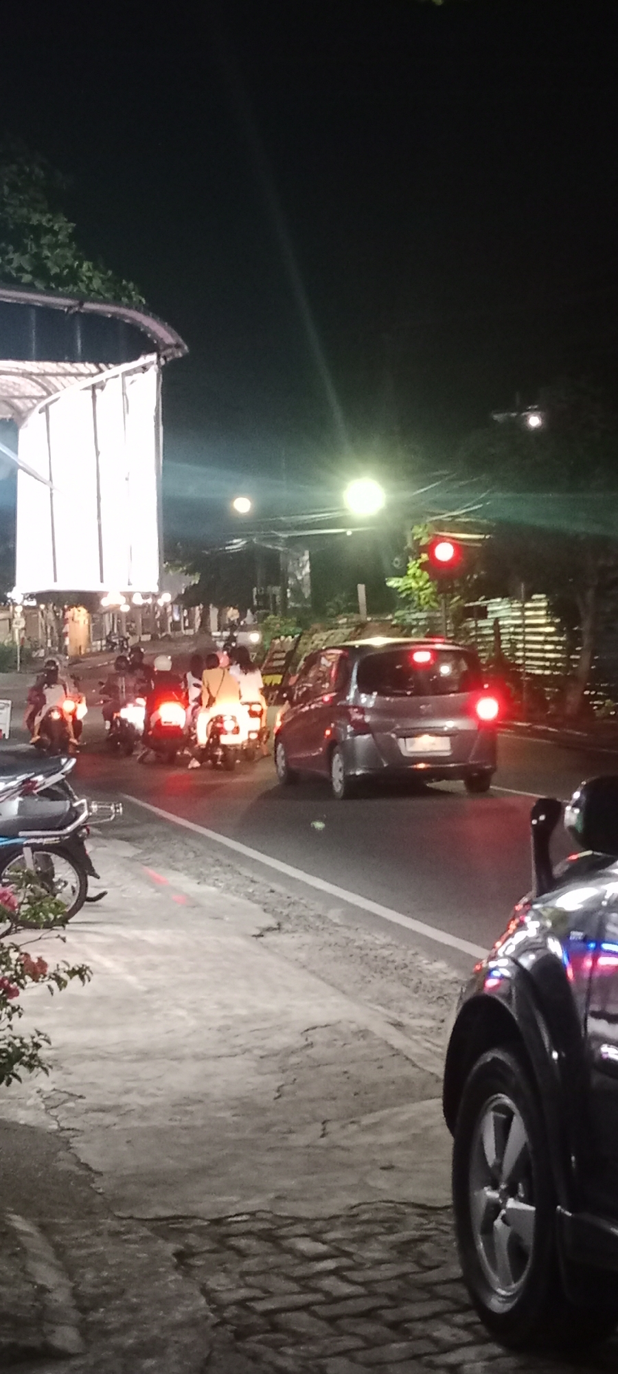 A traffic scene at night in Tanjung Pinang, Indonesia, features a row of motorcycles and a car stopped at a red traffic light. The motorcycles are mostly facing forward, with several riders visible. The car, a dark SUV, is positioned on the right side of the frame, also stopped. The road surface is asphalt with white lane markings. The surrounding environment includes a sidewalk made of textured material and brickwork, a building with illuminated panels on the left, and trees and other structures in the background. Streetlights and the traffic light itself provide illumination, casting reflections on the wet road surface.