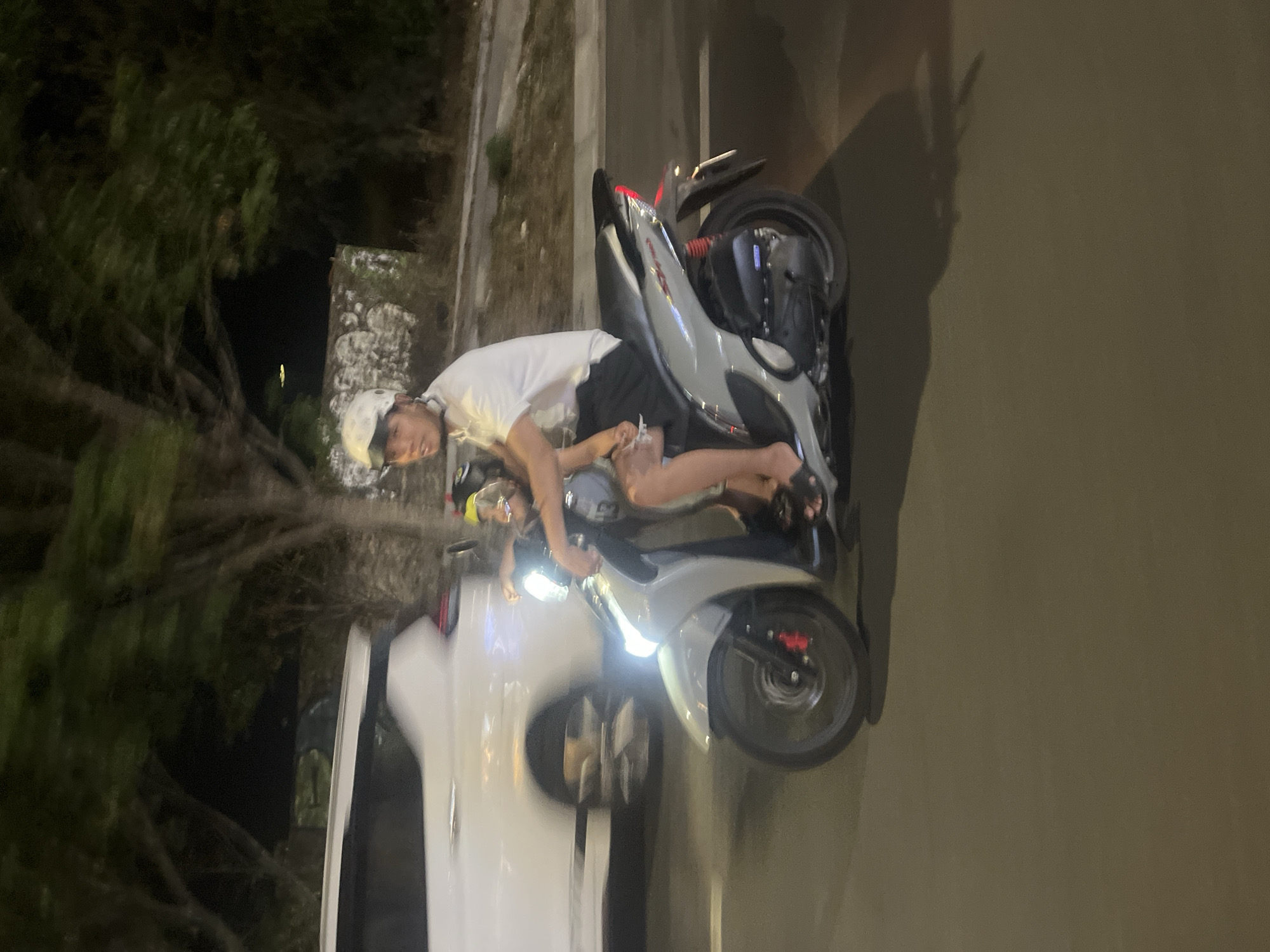 In the city of Đà Nẵng, Vietnam, at night, a person wearing a white helmet and a white t-shirt is riding a silver scooter. A child is seated in front of the rider. The rider is looking towards the left, and the child is looking forward. Both are dressed in casual clothing. A white vehicle is visible on the left side of the image, also in motion. The scene is illuminated by streetlights and the scooter's headlight. The background consists of trees and a stone wall.