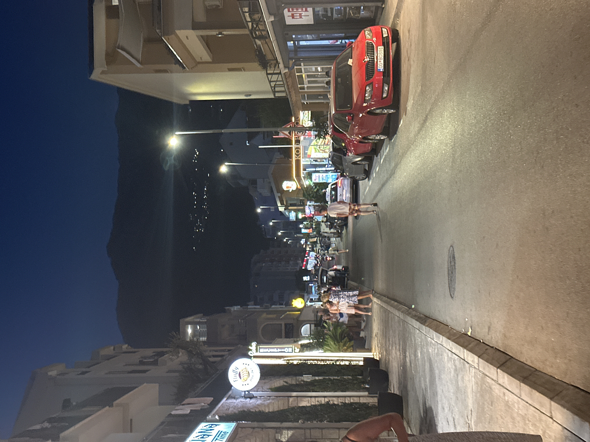 The image depicts a nighttime street scene in Budva, Montenegro. The street is lined with buildings, some of which appear to be shops or restaurants, indicated by lit signs and illuminated windows. Several cars are parked along the side of the road, and a few people are walking on the sidewalk and in the street. The sky is dark, suggesting it is late evening or night, and mountains are visible in the background, with lights from a distant settlement dotting the mountainside. Streetlights illuminate the scene, casting light on the road and the facades of the buildings. One building on the left has a blue neon sign that reads "lana time." Another sign above a shop features a stylized bread loaf. A red car with a license plate beginning with "OG BC" is prominently featured in the foreground on the right side of the road.