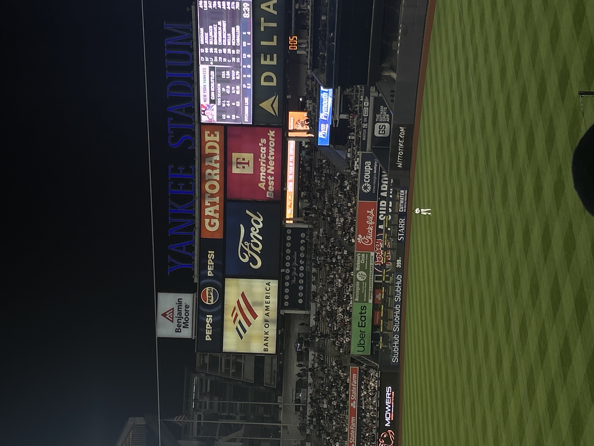 The image depicts Yankee Stadium in New York City at night. The stadium is largely filled with spectators. Prominent signage includes: "Yankee Stadium", advertisements for brands like Benjamin Moore, Pepsi, Gatorade, Ford, Bank of America, America's Best Network, Delta, Plymouth, State Farm, Uber Eats, Chick-fil-A, and Nitto Tire. The Yankee's lineup can be seen in the top left of the shot.