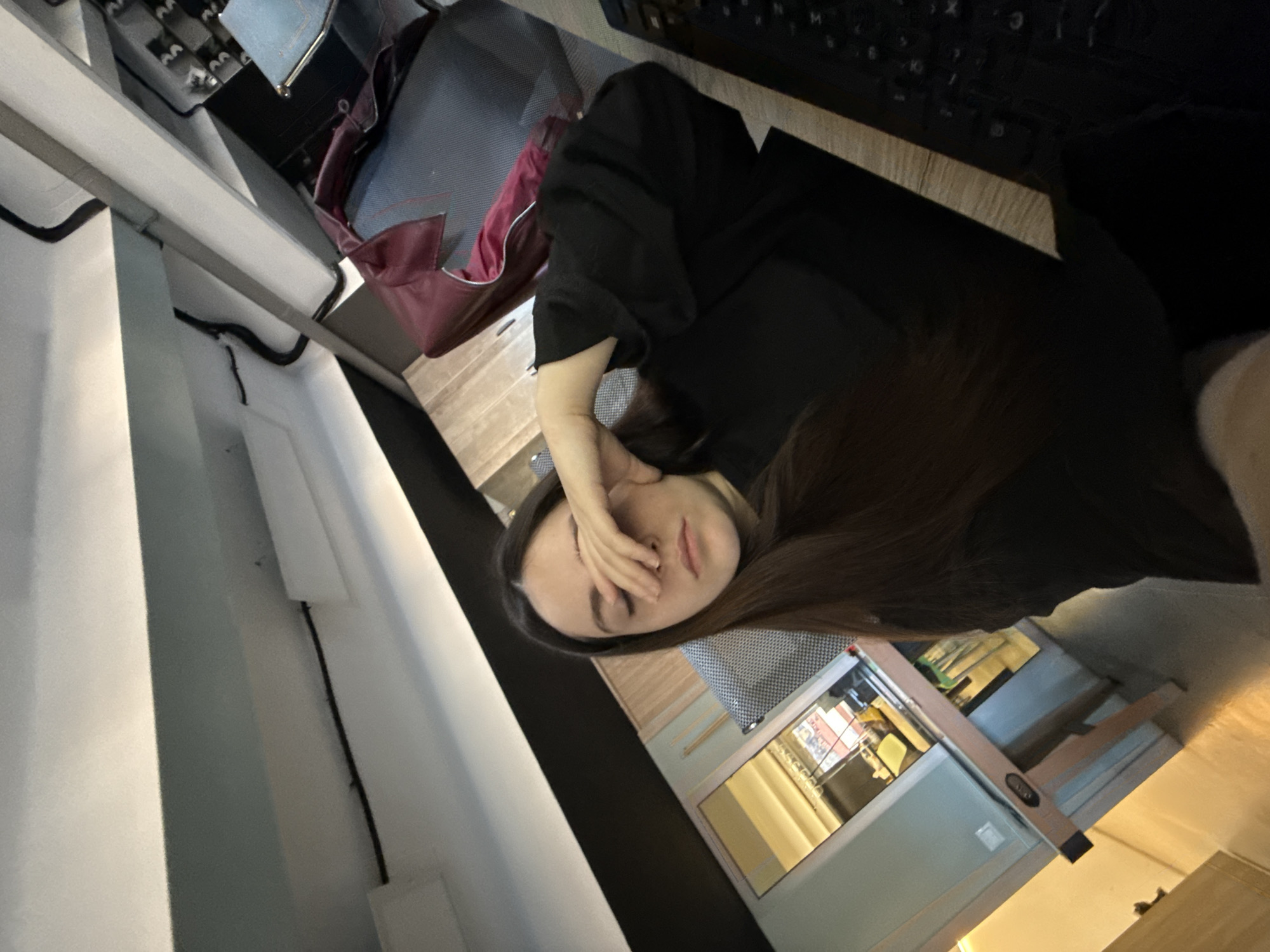 A woman is sitting at a desk next to a keyboard and possibly a chair. She has her face partially covered with her hand. The location is Yerevan, Armenia, as indicated by the furniture and objects present in the scene. There is also a display case or cabinet visible in the background. The scene appears to be indoors.
