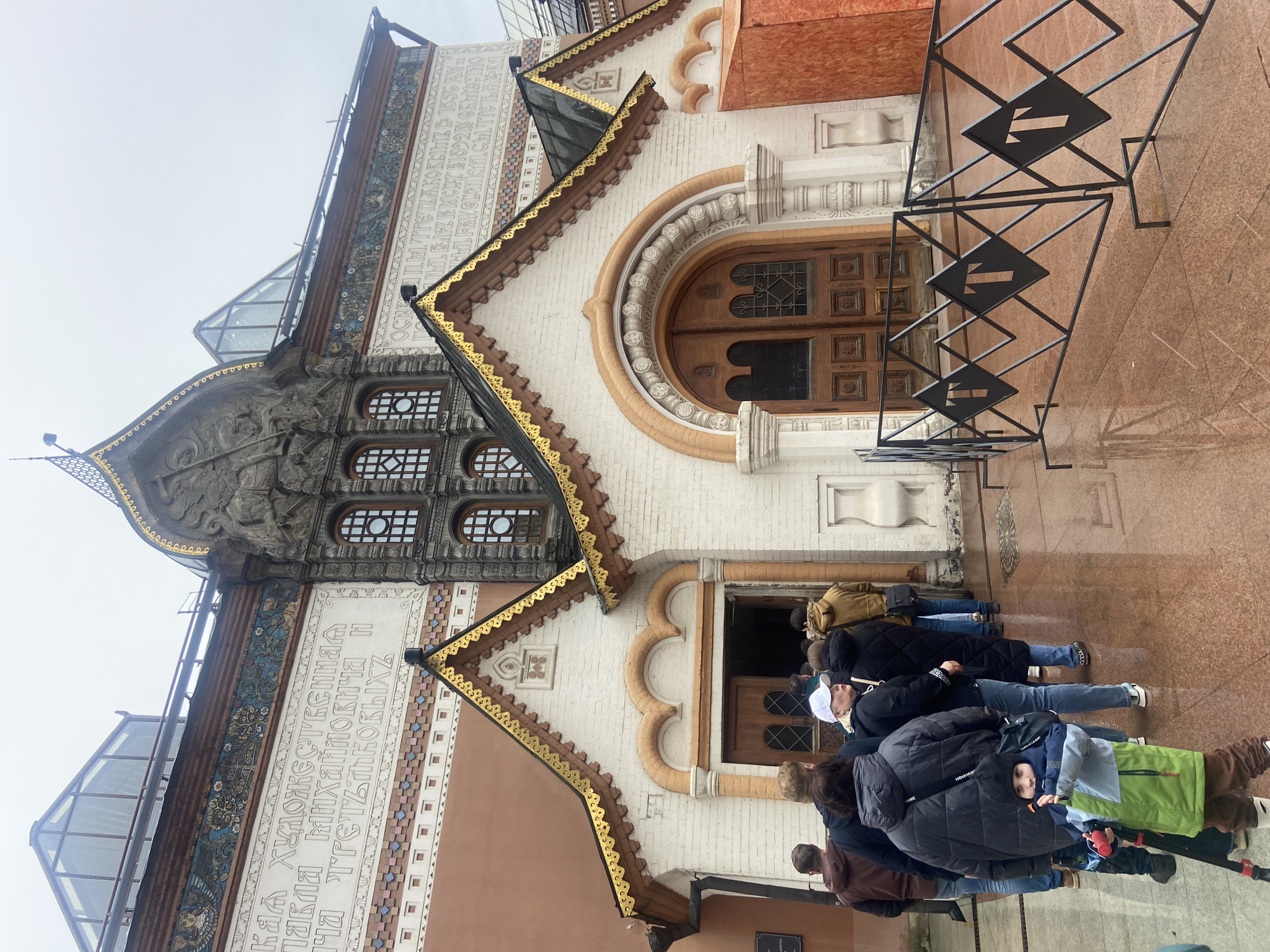 The image captures a daytime view of the State Tretyakov Gallery in Moscow, Russia. The architectural style features intricate detailing, including decorative inscriptions and ornate window frames. A line of people, primarily dressed in jackets, appears to be queuing to enter the building. One person stands out with a green and blue backpack. The overall environment suggests a cloudy day in an urban setting.
