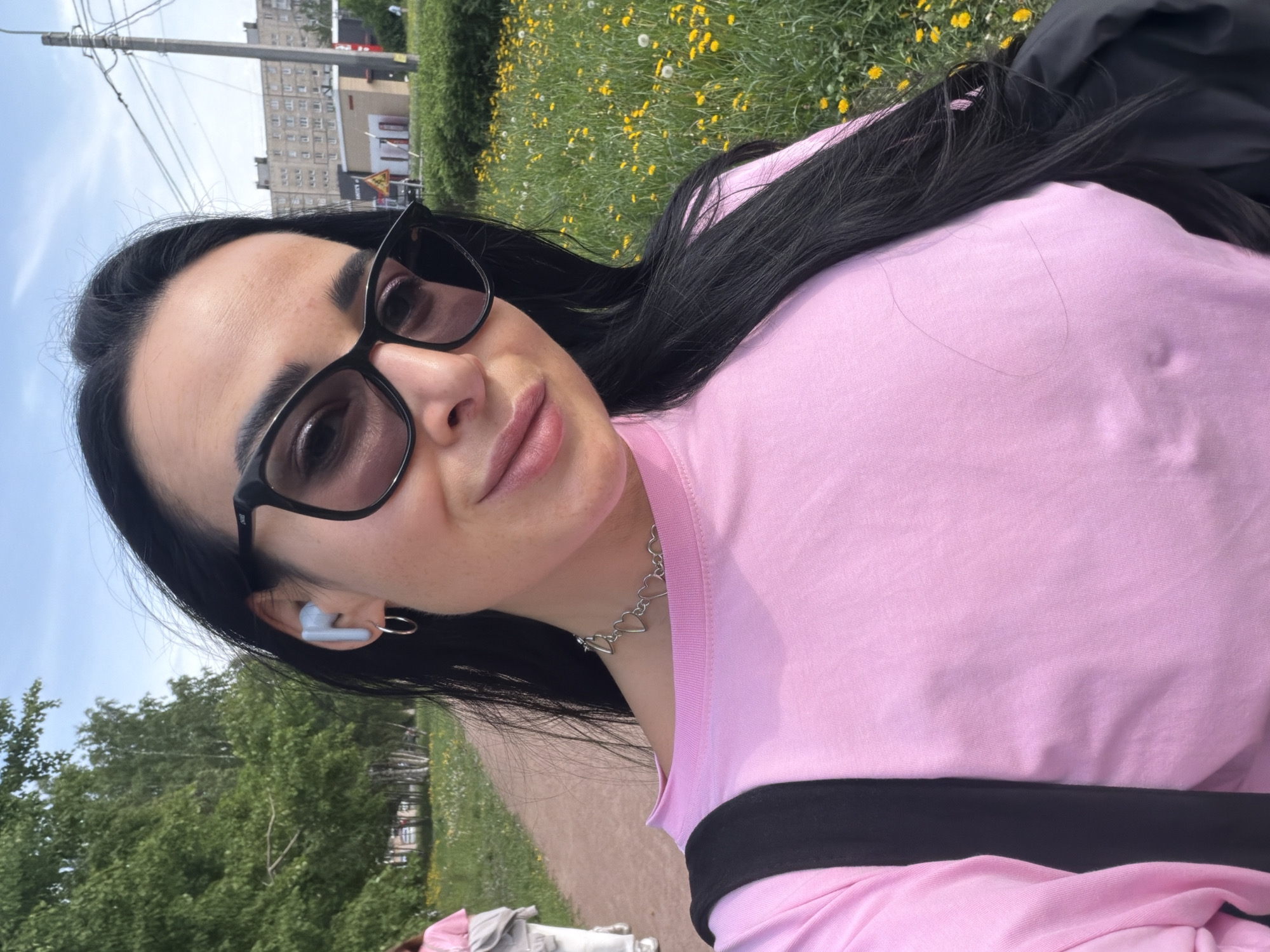 A person wearing sunglasses, a pink top, and a silver heart-shaped necklace is facing the camera. The person is wearing a blue wireless earbud in their left ear. A green field with yellow flowers is in the background. A cityscape with buildings is visible in the upper left corner. Based on architectural style and the overall environment, the location is Saint Petersburg, Russia.