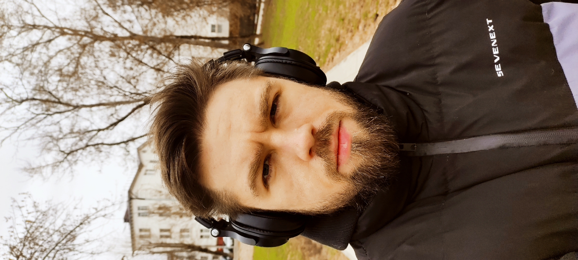 This image is a portrait-style photograph of a young man, taken outdoors in Fryazino, Russia. The man is positioned in the foreground, looking directly at the camera with a serious or contemplative expression, characterized by slightly furrowed brows and subtly parted lips. He has short brown hair, a full beard, and a mustache.
He is wearing black over-ear headphones and a black puffer jacket. The brand name "SEVENEXT" is visible in white lettering on the right shoulder area of his jacket.
The background suggests a park or urban green space during a late autumn or winter day. Bare tree branches are prominent against an overcast sky, and hints of light green grass and a paved path are visible. In the distance, residential buildings with visible windows can be seen through the sparse trees. The lighting is soft and diffused, indicating an overcast day. The scene captures a casual moment of the man outdoors, possibly listening to music or a podcast.