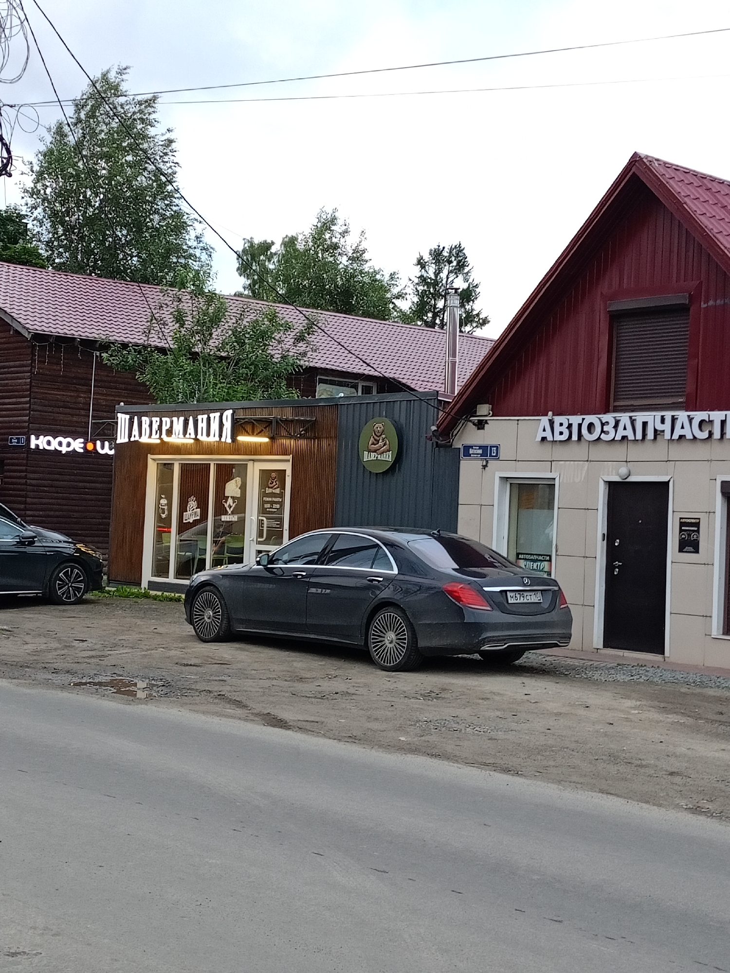 A dark-colored sedan is parked on a dirt lot next to a paved road. In the background, there are several buildings with red roofs. One building has a sign that reads "АВТОЗАПЧАСТИ" (Autoparts) in large white letters. Another building has a sign that reads "ШАВЕРМАНИЯ" (Shawarma) in illuminated white letters. There are trees and overhead wires visible in the scene. The sky is overcast. The car's license plate is visible and reads "M679CT 190".