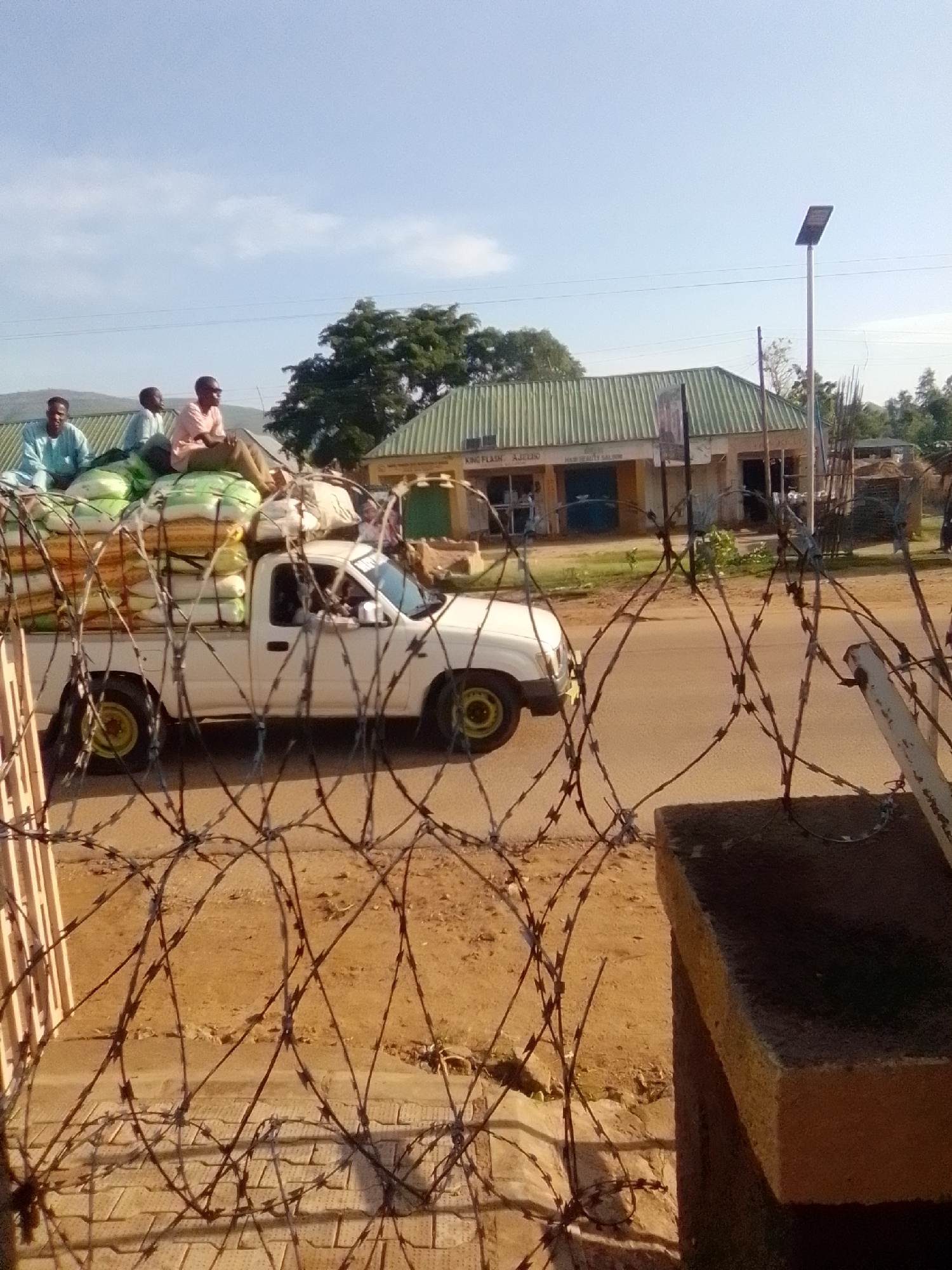 In Galadima, Nigeria, a white pickup truck loaded with sacks of goods, likely agricultural produce, is parked on a dirt road in front of a building with a green corrugated metal roof. Three men are seated on top of the sacks in the truck bed. The truck is partially obscured by a fence made of barbed wire. The building behind the truck appears to be a commercial establishment, with signage visible on its facade, including "KING FLASH AJILELO" and "HAIR BEAUTY SALON." A solar-powered street lamp is visible to the right of the building. The sky is clear with some scattered clouds.