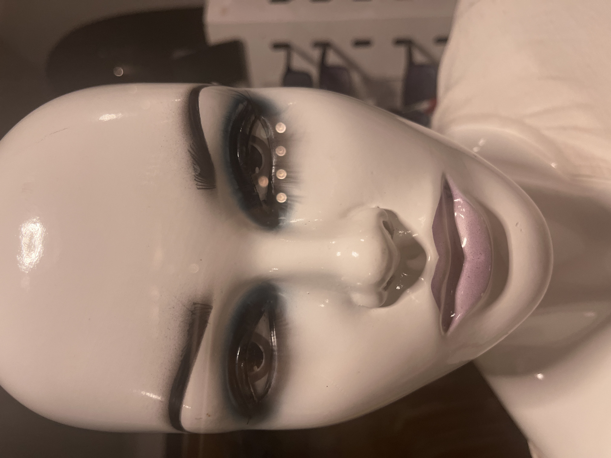 A close-up, slightly off-kilter image depicts the head of a mannequin. The mannequin's skin is a glossy white, and it has painted-on features. The eyes are detailed with black eyeliner and eyeshadow, and one eye has a row of small, reflective dots along the upper lid. The lips are painted in a shade of light purple. The background is blurred and includes hints of other objects, including what appears to be a black surface and some indistinct shapes. The environment suggests an indoor setting, potentially a store or studio where mannequins are used. There is no indication of the location of Budva, Montenegro in the image.