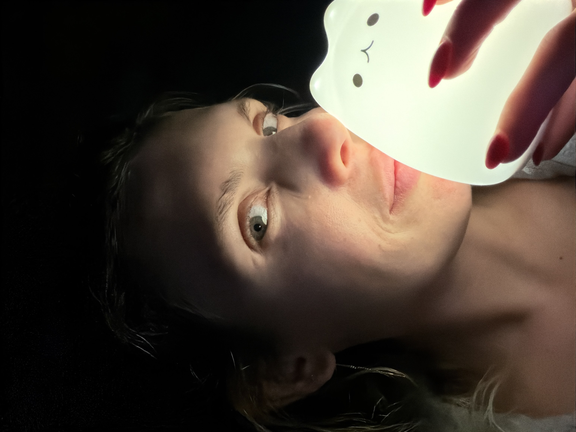 A close-up, vertically oriented image captures a young woman's face partially illuminated by a glowing, white, cat-shaped night light. The woman's face is seen from the nose upwards, with her right eye, eyebrows, and forehead visible and softly lit by the upward glow. Her light-colored eye is open and appears to be looking slightly upwards, possibly at the light itself or beyond it. The cat-shaped light, featuring simple black dots for eyes and a small curved line for a mouth, is held by a hand with red-painted fingernails, positioned below and to the right of the woman's face. The light strongly illuminates the underside of her chin, lips, and nose, creating a dramatic and somewhat playful lighting effect against the otherwise dark background. The scene is enveloped in near darkness, emphasizing the solitary light source and the contours of her face and hand. This suggests a nighttime or very dimly lit indoor setting, possibly in Budva, Montenegro, though no specific location cues are visible within the frame. There is no visible text in the image.