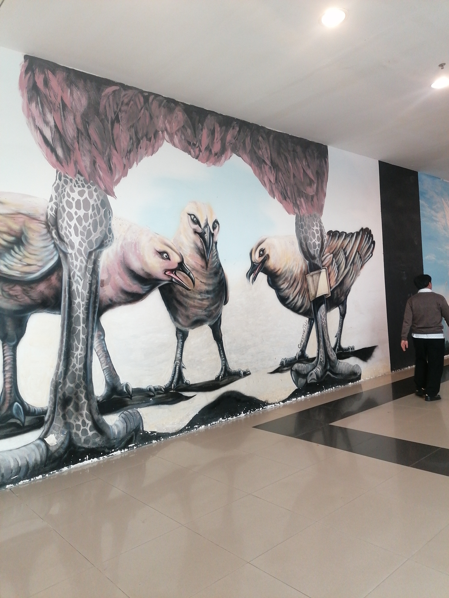 This image presents an indoor hallway in Da Lat, Vietnam, dominated by a large mural on the left wall. The mural depicts four fantastical, oversized, bird-like creatures rendered with a trompe l'oeil effect, giving them a three-dimensional appearance. These creatures possess light-colored bodies, distinct dark beaks, and prominent, intense eyes, standing on thick, dark, textured legs and feet that cast painted shadows on the floor. An archway of dark reddish-brown, feather-like structures frames the upper portion of the mural, while grey and white patterned column-like elements appear to separate the creatures. A signature reading "CINESTAR D..." is visible on the right leg of the second creature from the right. The background behind the figures is a light blue and white, suggesting a sky. To the right of the mural, the wall transitions to a solid black section before a faint blue and white sky mural becomes visible. The floor is covered with polished, light beige square tiles, interrupted by a sharp diagonal pattern of dark black rectangular tiles extending from the foreground to the right. The white ceiling features several recessed circular lights. On the far right of the scene, a person with dark hair, dressed in a dark brown long-sleeved top, a white collar, and black trousers, walks away from the viewer, heading towards the right along the tiled floor.