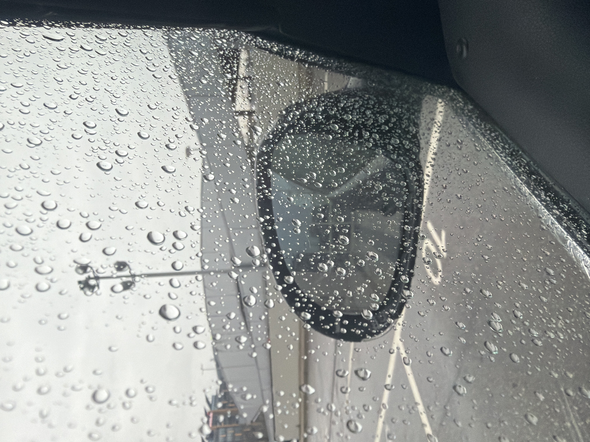 The image shows a view from inside a vehicle, likely a car, looking out through a rain-covered window. The focus is on the raindrops clinging to the glass. Visible through the blurred and distorted raindrops are hints of the outside environment. There's what appears to be a side mirror of the vehicle, also covered in raindrops, which partially reflects the outside scene. A blurred structure with vertical lines is noticeable, possibly a building or bridge. Also visible are what appears to be street lights.
The city of Sofia, Bulgaria is not specifically identifiable from this image.