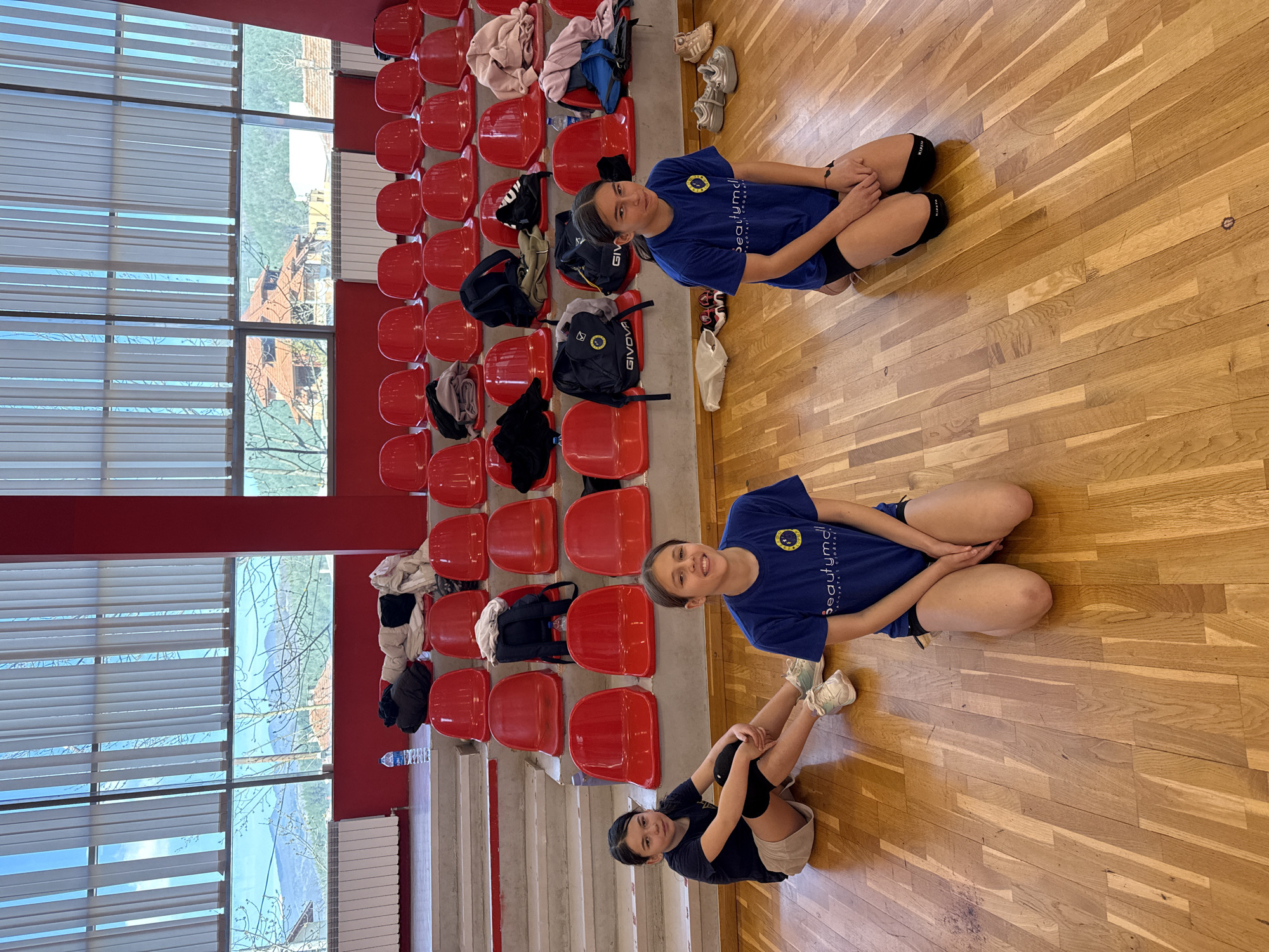 Three adolescent females are present in an indoor gymnasium setting. The room features wooden flooring and red spectator seating. The seating rows on the left side of the image are partly occupied with bags and clothes. The leftmost section includes stairs leading up to the seating area and a large window with blinds. Outside the window, a partial view of a lake and structures possibly located in Etropole, Bulgaria can be seen. The two girls in the upper half of the frame are wearing matching blue sports shirts with a logo of the “Beautymold” company and are kneeling on the floor. The third girl is in the lower part of the image. She is kneeling with her knees up. She is wearing a black t-shirt.