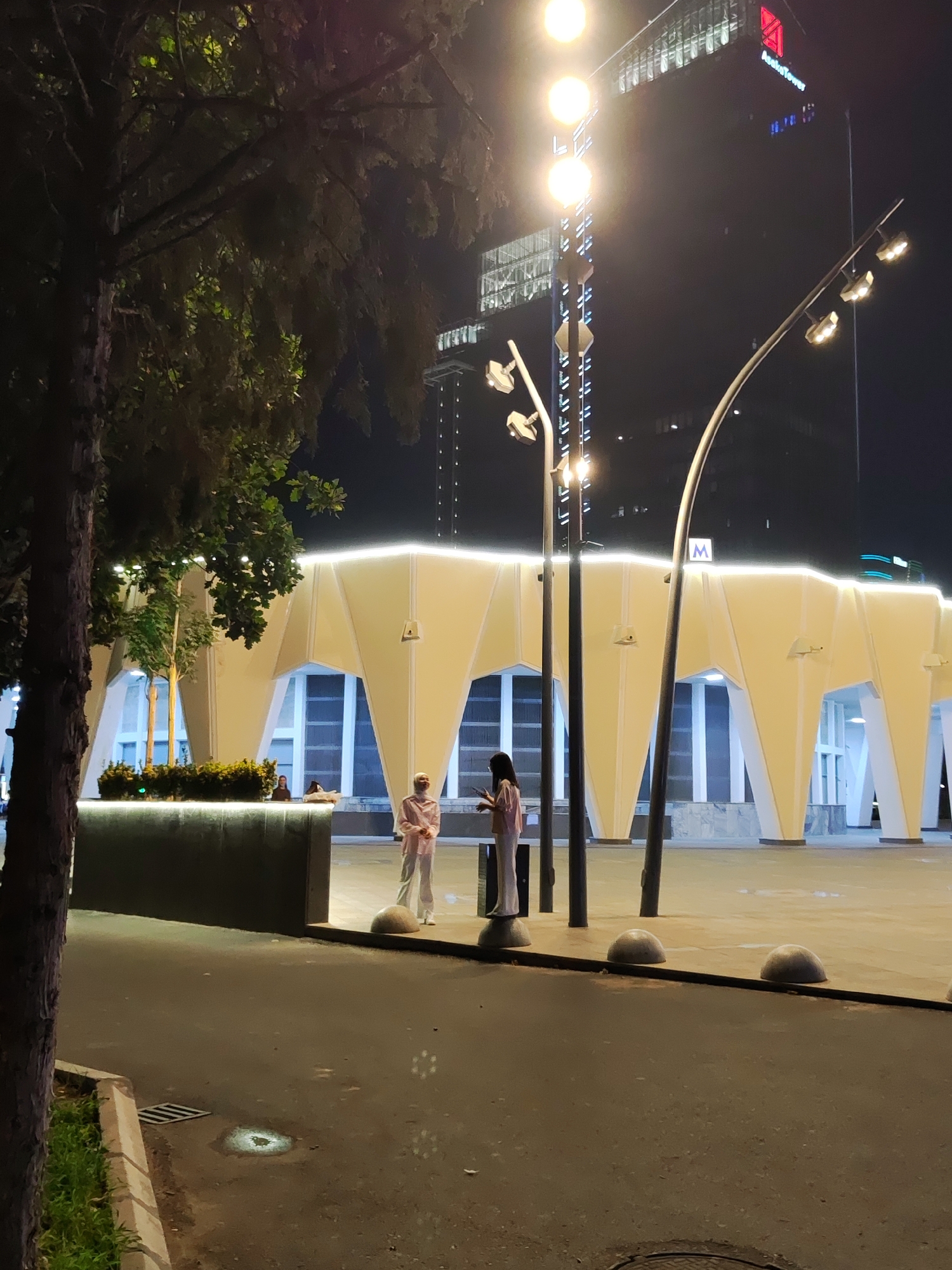 The image shows a nighttime scene in Tashkent, Uzbekistan, with the "Anhor Tower" building visible in the background. The foreground features a modern, illuminated structure with large, angular supports and a bright, white light strip along its top edge. Two individuals are standing in front of this structure. One, on the left, wears a pink outfit and stands facing right, looking at the other person. The second individual, on the right, is dressed in a light-colored, loose-fitting outfit and is looking down at their phone. A tree with dark branches and green foliage is to the left of the frame, partially obscuring the view of the building. Several streetlights with multiple bulbs are also present, illuminating the area. The ground in the foreground is paved, with a sidewalk and some rounded concrete bollards. The scene appears to be an urban public space.