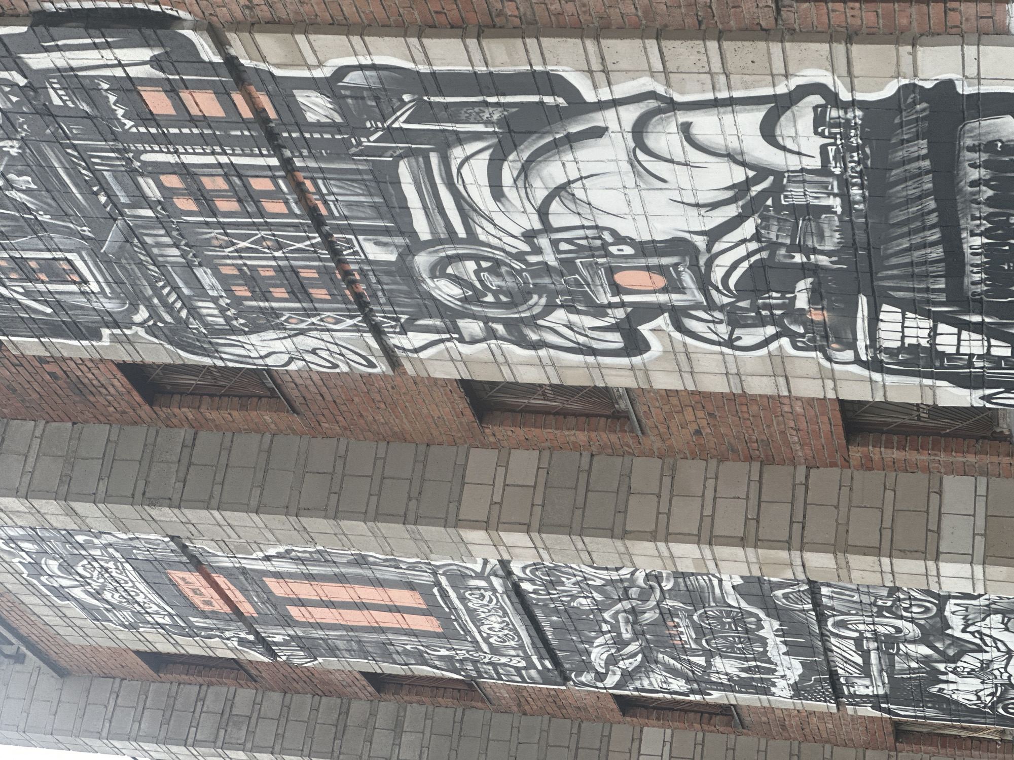 The image shows the underside of a bridge in Novosibirsk, Russia. The bridge is constructed with alternating rows of red brick and grey brick blocks. The underside is decorated with black and white graffiti art depicting architectural elements like windows and building facades, along with abstract swirling shapes. The style is urban and somewhat stylized.