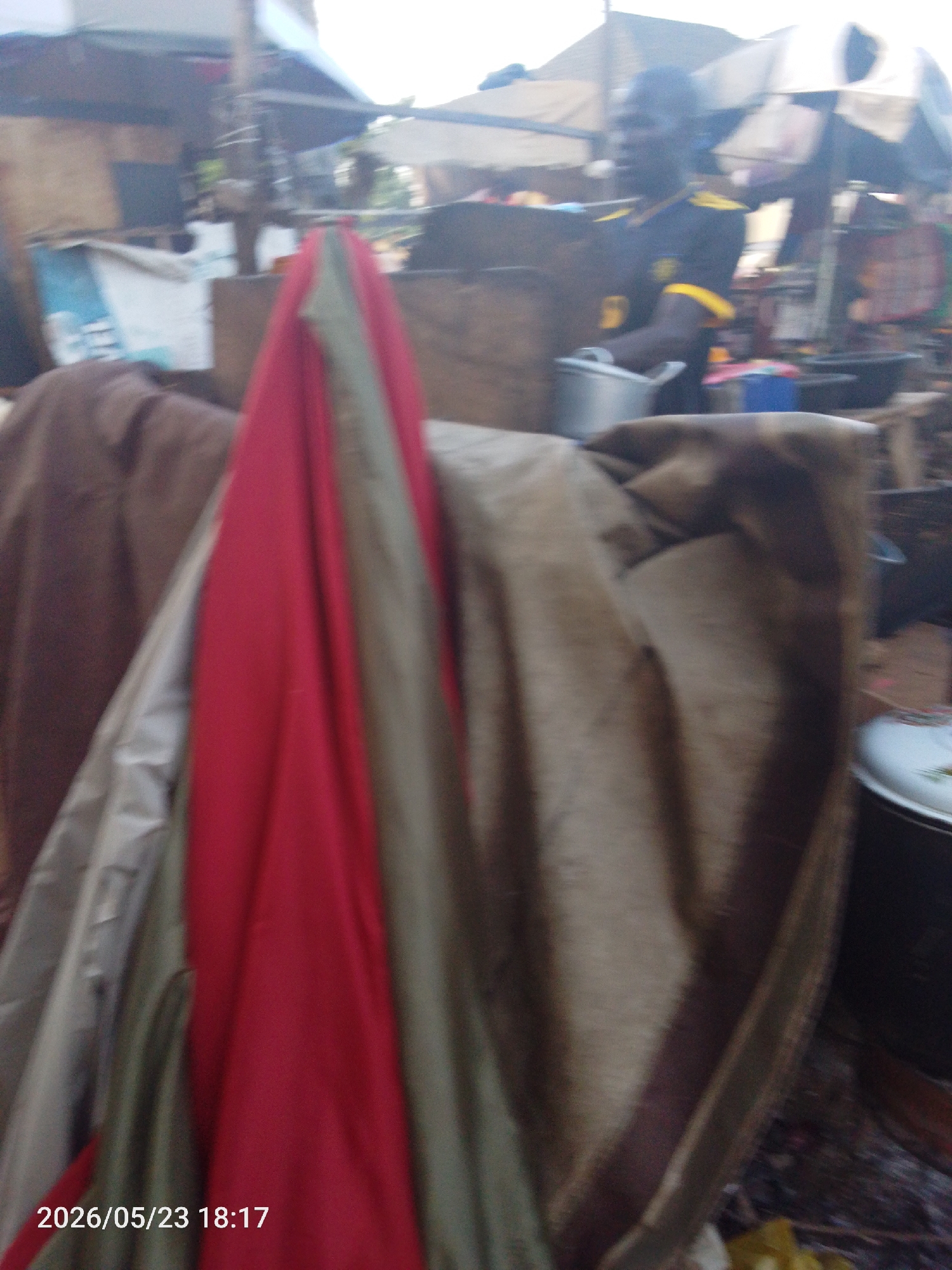 The image depicts an outdoor scene, possibly a market or a street food area, in Lokoja, Nigeria. A man wearing a dark shirt with yellow accents is seen holding a metal pot. Various stalls and tents are visible in the background. In the foreground, there are draped fabrics in shades of red, grey, and brown. There are cooking pots nearby.