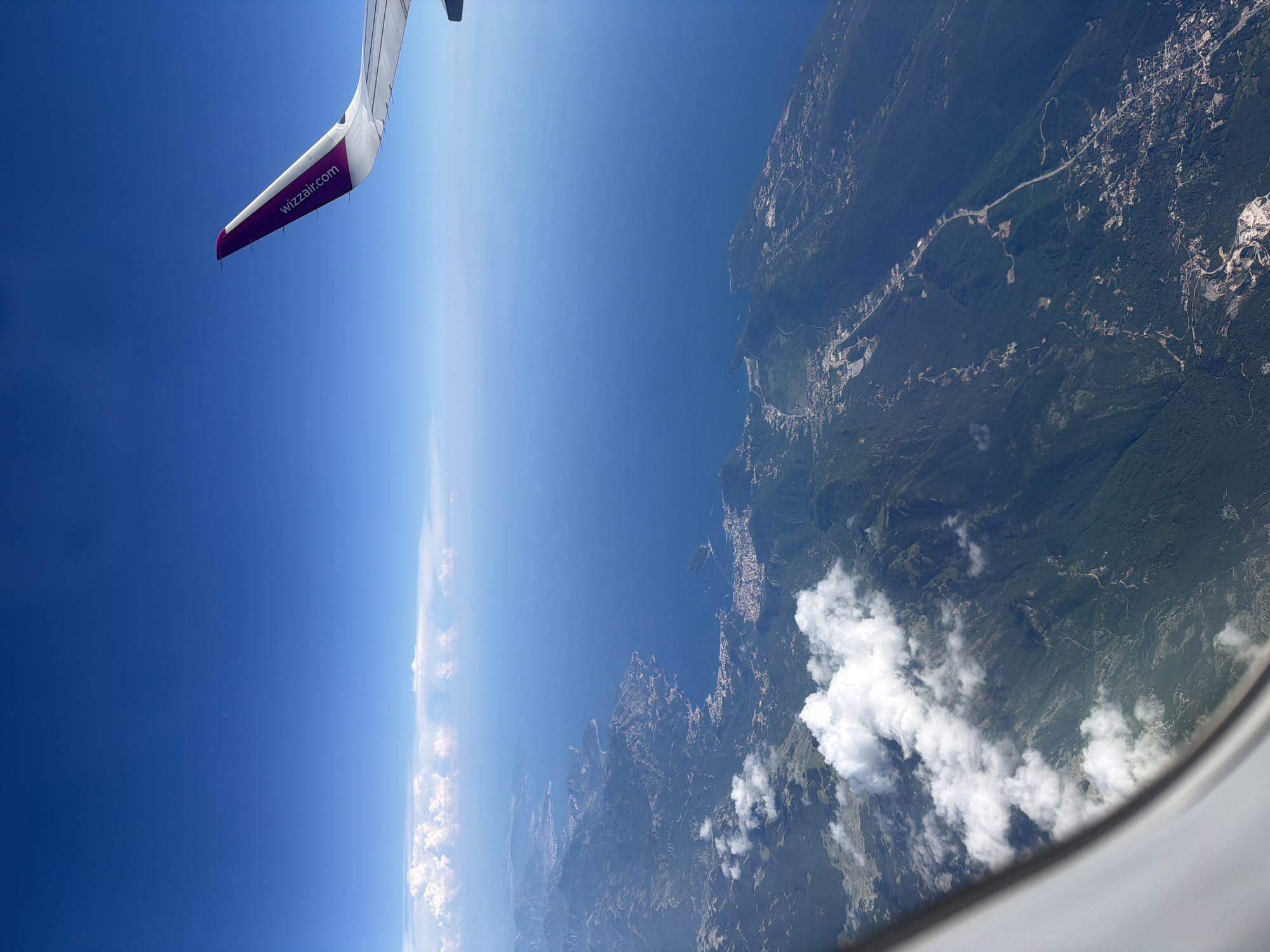 An aerial view from an airplane window shows a coastline with lush green mountains meeting the deep blue sea. A prominent airplane wing, with the Wizzair.com logo in purple, extends into the upper right corner of the frame. White clouds are scattered both above and below the aircraft, with a thin line of clouds visible on the horizon where the sky meets the sea. The coastline is dotted with small towns and settlements, with roads winding through the mountainous terrain and along the edge of the water. The city of Donja Cijevna, Montenegro, is visible along the coast, characterized by its dense development nestled between the mountains and the Adriatic Sea.