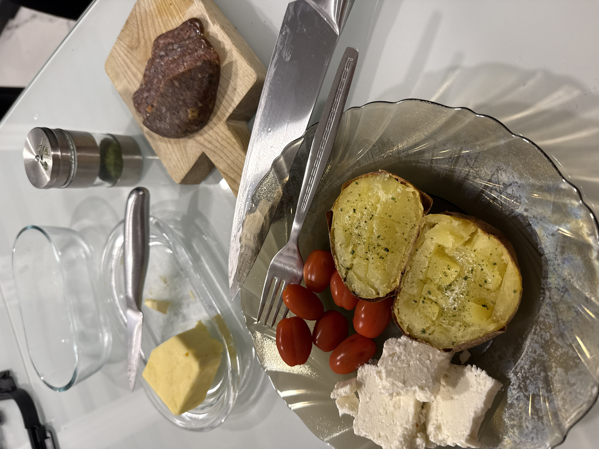 The image depicts a table setting with a glass tabletop. A light-colored, presumably wooden, cutting board holds a piece of salami. To the left of the salami is a glass container with a metallic top. Adjacent to this, there is a small dish containing a cube of butter, accompanied by a knife. The main plate is transparent with a decorated edge, featuring two halves of baked potatoes, some cherry tomatoes, and several pieces of white cheese. A knife and fork are placed on the plate. The setting gives off a minimalistic, modern aesthetic. There is no information available in the image to confirm the city of Burgas, Bulgaria.