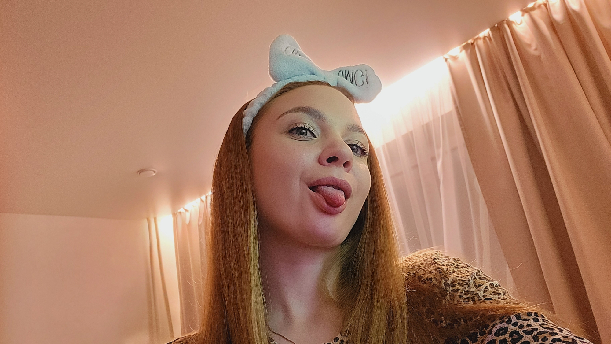 A young woman with light brown hair and a blue bow headband is making a silly face with her tongue sticking out. She is wearing a leopard print shirt. Behind her is a room with beige curtains and what appears to be recessed lighting. Based on user geolocation, the location is Chaikovsky, Russia.
