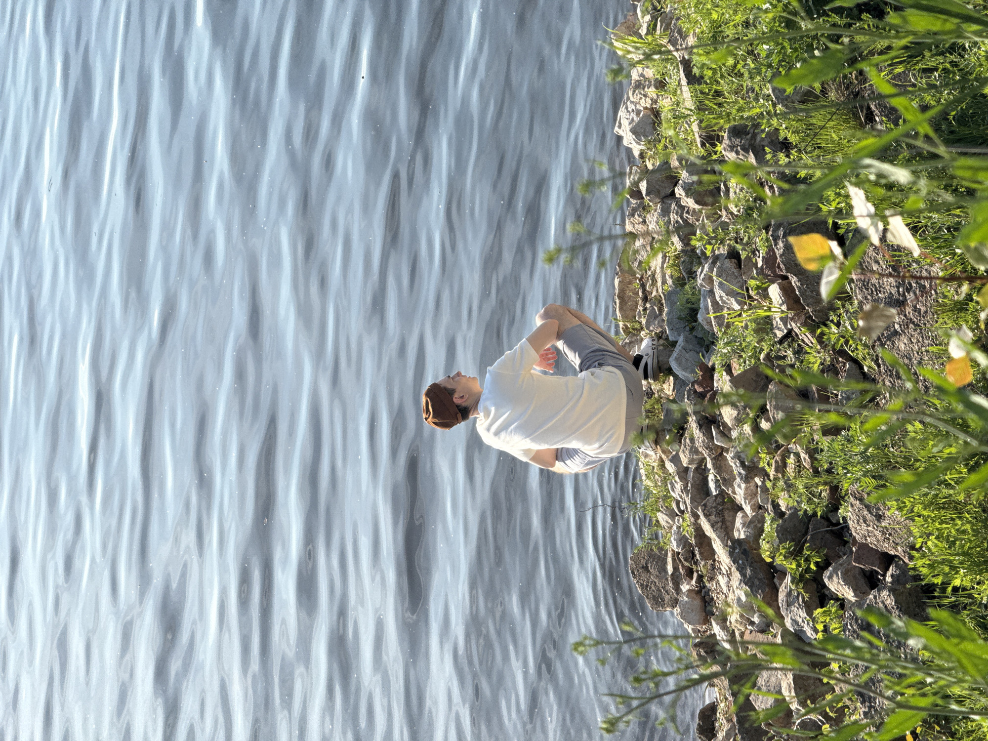 A young man sits on a rocky shoreline alongside a body of water, likely a river or lake, in what is presumably Chaikovsky, Russia. He is wearing a white t-shirt, grey shorts, and a brown head covering. He is sitting with his legs bent and his back towards the water. The water is light blue-grey with a rippled surface. The shoreline consists of various-sized grey rocks interspersed with green grass and foliage.