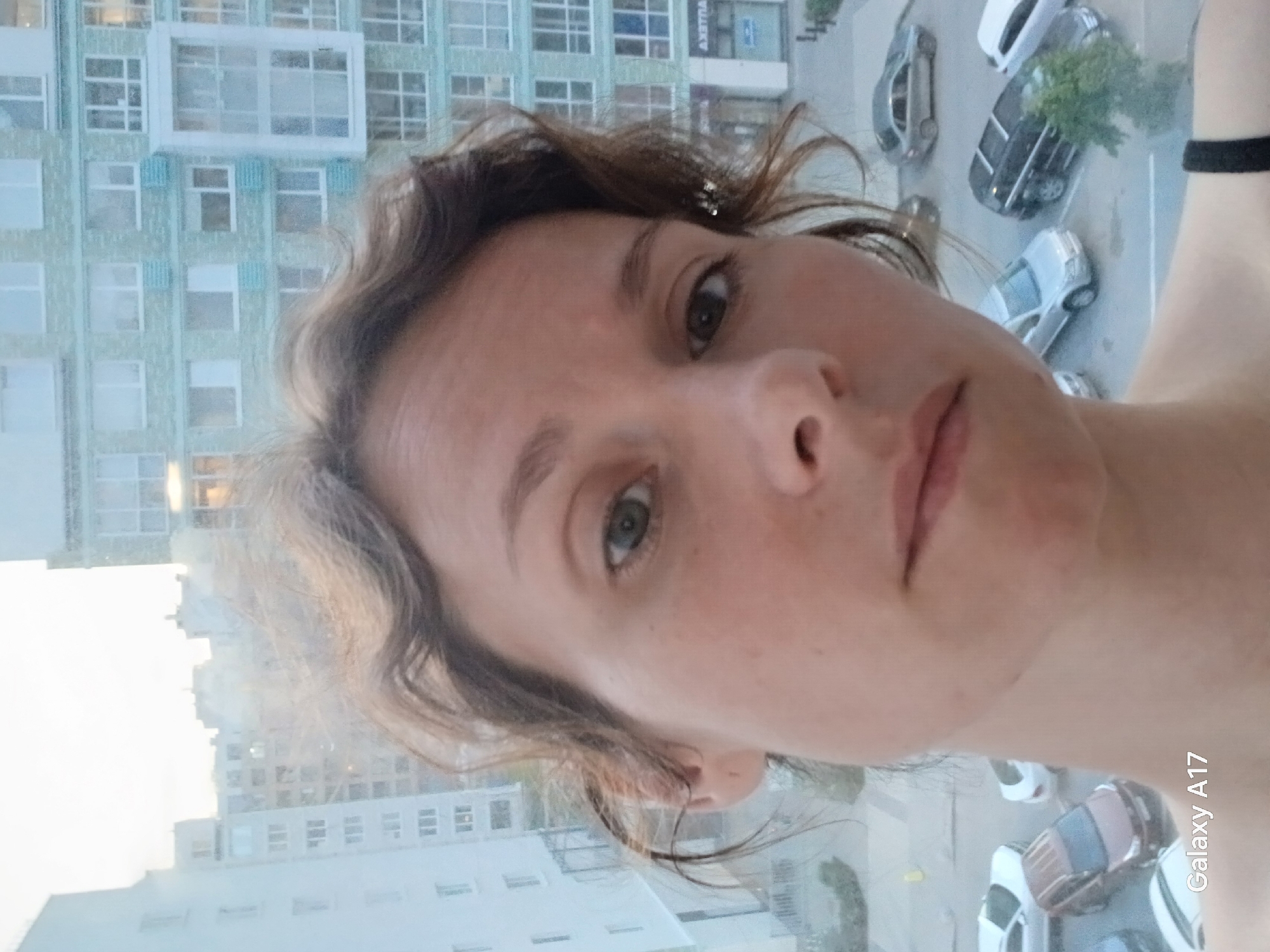 A woman takes a selfie from a high vantage point, likely a balcony or tall building. The background shows a cityscape with modern apartment buildings. Cars are parked on the street below. The image was taken with a Samsung Galaxy A17. The image does not allow for positive identification of a specific location within Yekaterinburg.