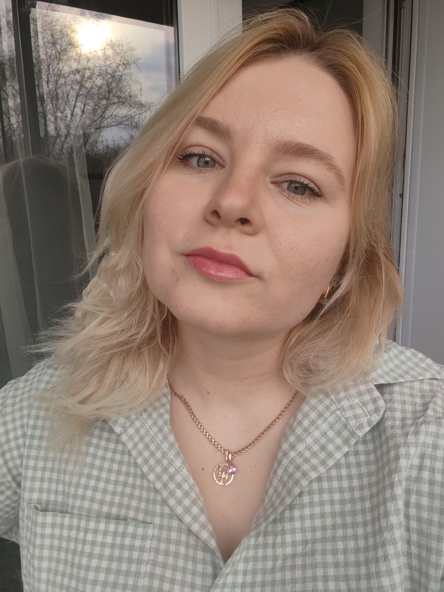 A close-up shot depicts a fair-skinned woman with blonde, shoulder-length hair. She has light eyes and her lips are coated in a glossy pink lipstick. She wears a light green and white checkered shirt and a gold necklace with a pendant containing a pink gemstone. The background shows a window with reflections of trees and sunlight, likely indicating an outdoor setting. There is no verifiable information to ascertain that the location is in the city of Izhevsk, Russia.