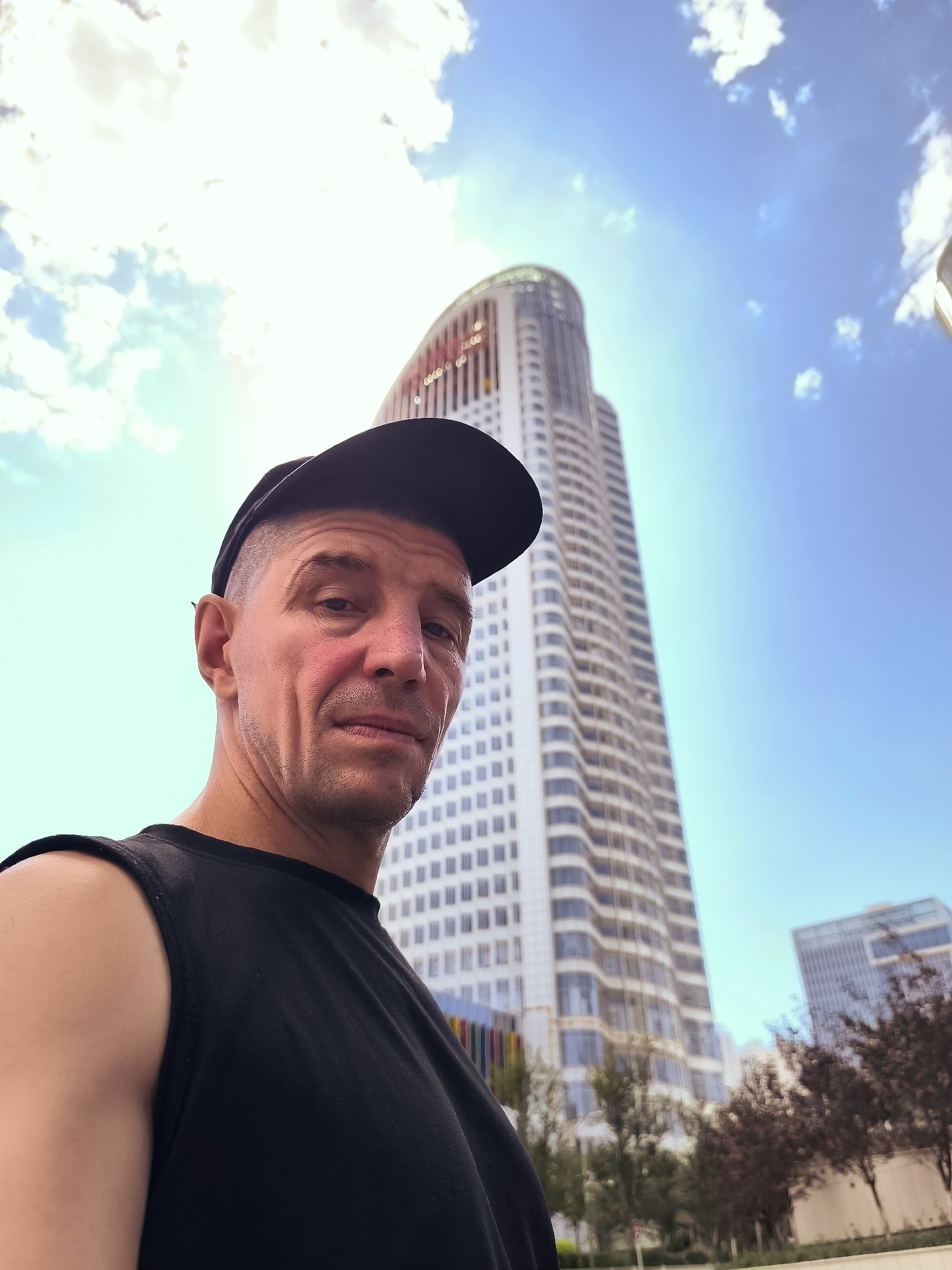 The image is a low-angle, outdoor, selfie-style shot featuring a man in the foreground wearing a black sleeveless shirt and a black baseball cap. He has fair skin and appears to be in his late 40s or 50s, with lines visible on his face and a slight flush on his cheeks. His expression is neutral, and he is looking directly at the camera. Behind him, a tall, modern skyscraper with a curved top dominates the background. The building is white with numerous windows. To the right of the main skyscraper, a second, slightly shorter building is visible, also appearing to be modern. The sky is bright blue with scattered white clouds, and the sun is positioned high, creating a lens flare in the upper right quadrant of the image. Trees with dark red foliage are scattered in front of the buildings. The overall environment suggests an urban setting.