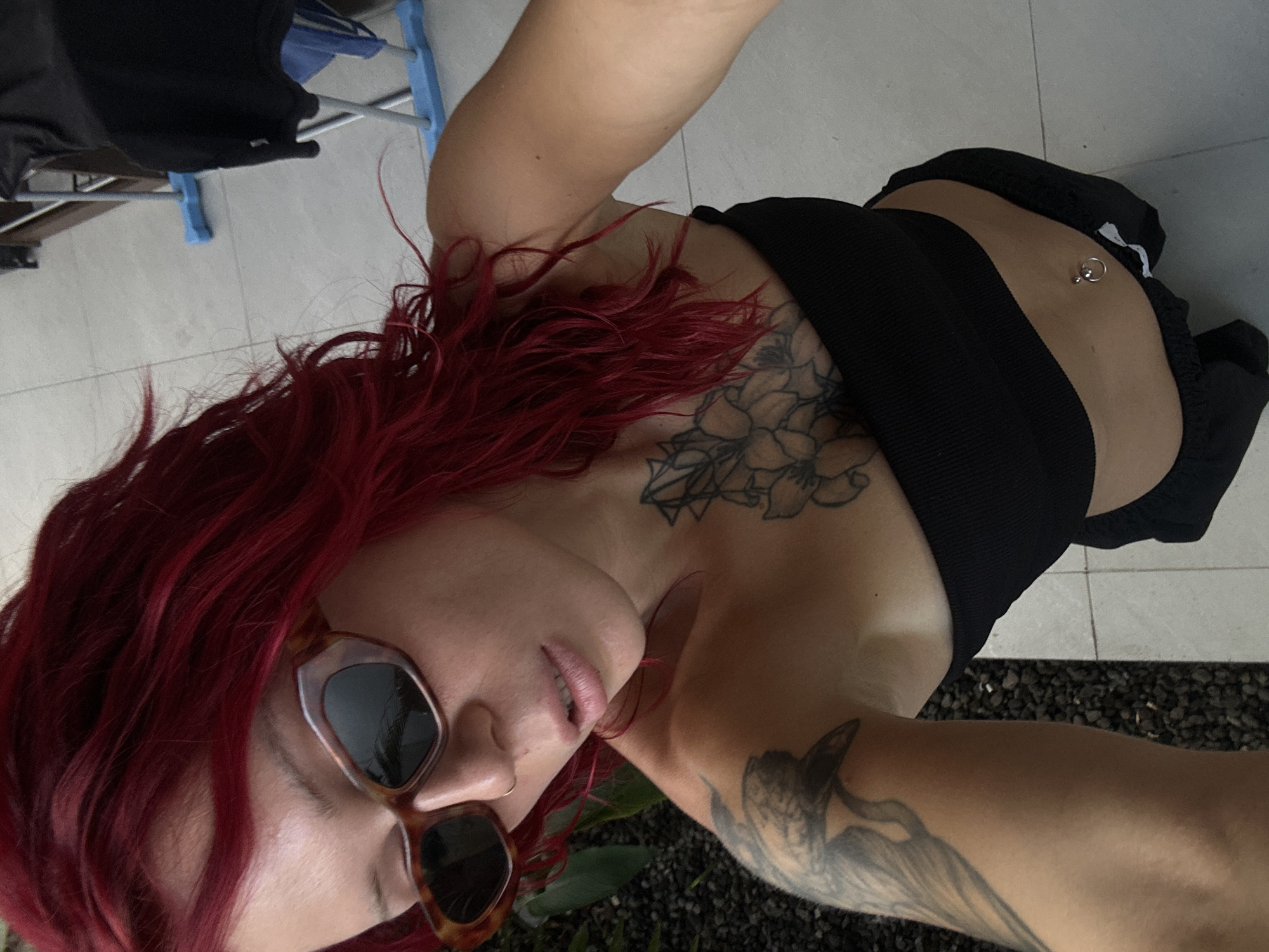 A close-up selfie captures a person with vibrant red, curly hair, lying down outdoors, looking upwards into the camera. They wear dark, tortoise-shell framed sunglasses that obscure their eyes, and their lips are slightly parted. A subtle nose piercing is visible.
The individual is adorned with several tattoos: a large floral design covers part of their chest and shoulder, and a bird-like figure is visible on their right upper arm. They are wearing a black, ribbed crop top or bikini top, revealing a navel piercing. Their skin appears tanned.
The setting suggests an outdoor or semi-outdoor space in North Kuta, Indonesia. The background includes light-colored tiled flooring and a section of dark, possibly gravelly or pebbled, ground. To the upper left, a blue and white clothes drying rack is partially visible, with a black garment hanging on it. The lighting is bright, indicating daytime. The person's relaxed posture and closed eyes behind the sunglasses convey a sense of leisure and calm. No visible text is present in the image.