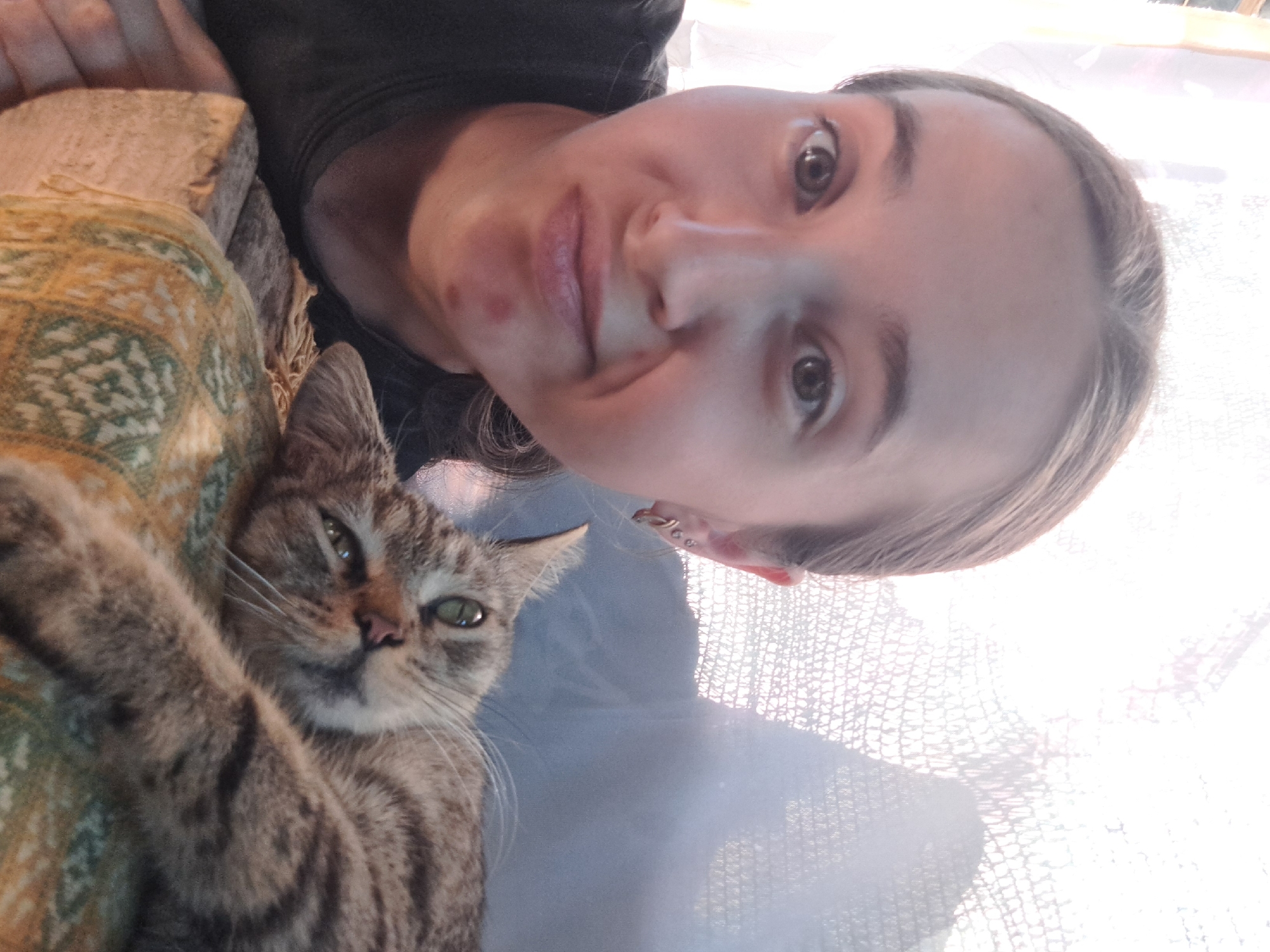 The image is a close-up selfie of a young woman and a tabby cat. The woman is in the foreground, looking at the camera with a slightly puzzled expression. She has light brown hair pulled back and is wearing a dark t-shirt. A red pimple is visible on her chin. Next to her, the cat is lying down, its head resting on a patterned fabric. The cat has green eyes and is looking slightly away from the camera. The background is blurred, showing what appears to be a window with diffused light and a textured surface, possibly a curtain or net. The overall impression is a candid, casual moment captured indoors. The location of Vypasne, Ukraine, is not evident from the image itself.