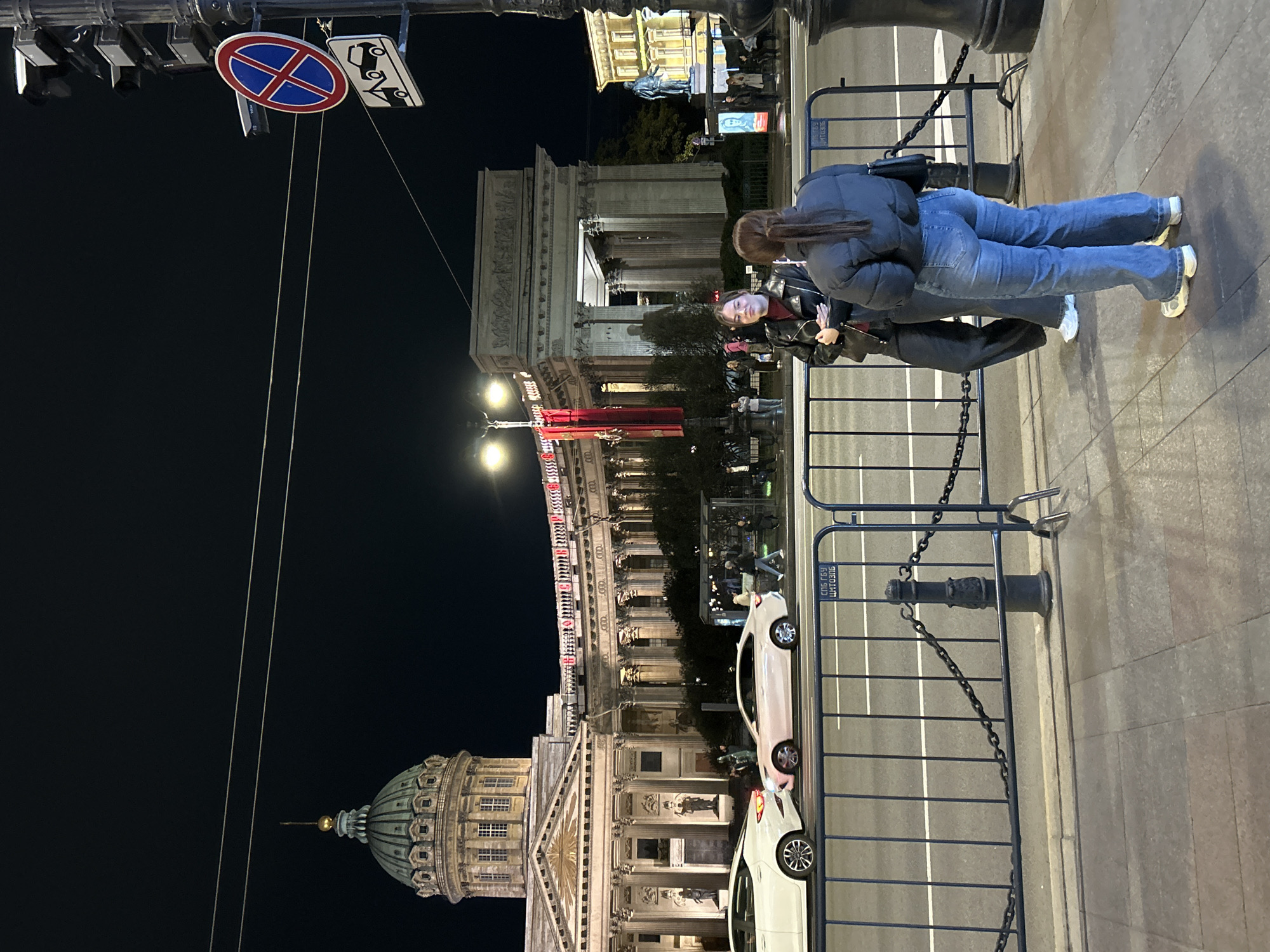 Two individuals stand on a sidewalk, near metal barriers, with vehicles visible in the roadway. In the background stands the Kazan Cathedral, adjacent to the Nevsky Prospect (street with buildings). Above, signage includes "no parking" and "tow away zone". The image was captured at night.