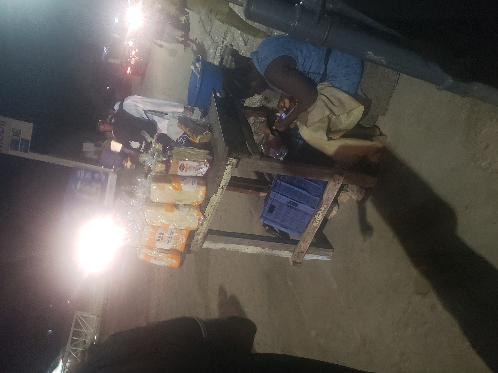 The image shows a street scene at night in Alalubosa, Nigeria. A woman is crouched near a wooden table on which several loaves of bread are stacked. She appears to be interacting with a mobile device, possibly making a transaction or communicating. A blue plastic crate sits underneath the table. To the left, a Mobil petrol station sign is visible, and cars are driving past. The scene is illuminated by streetlights and headlights, creating a high contrast.