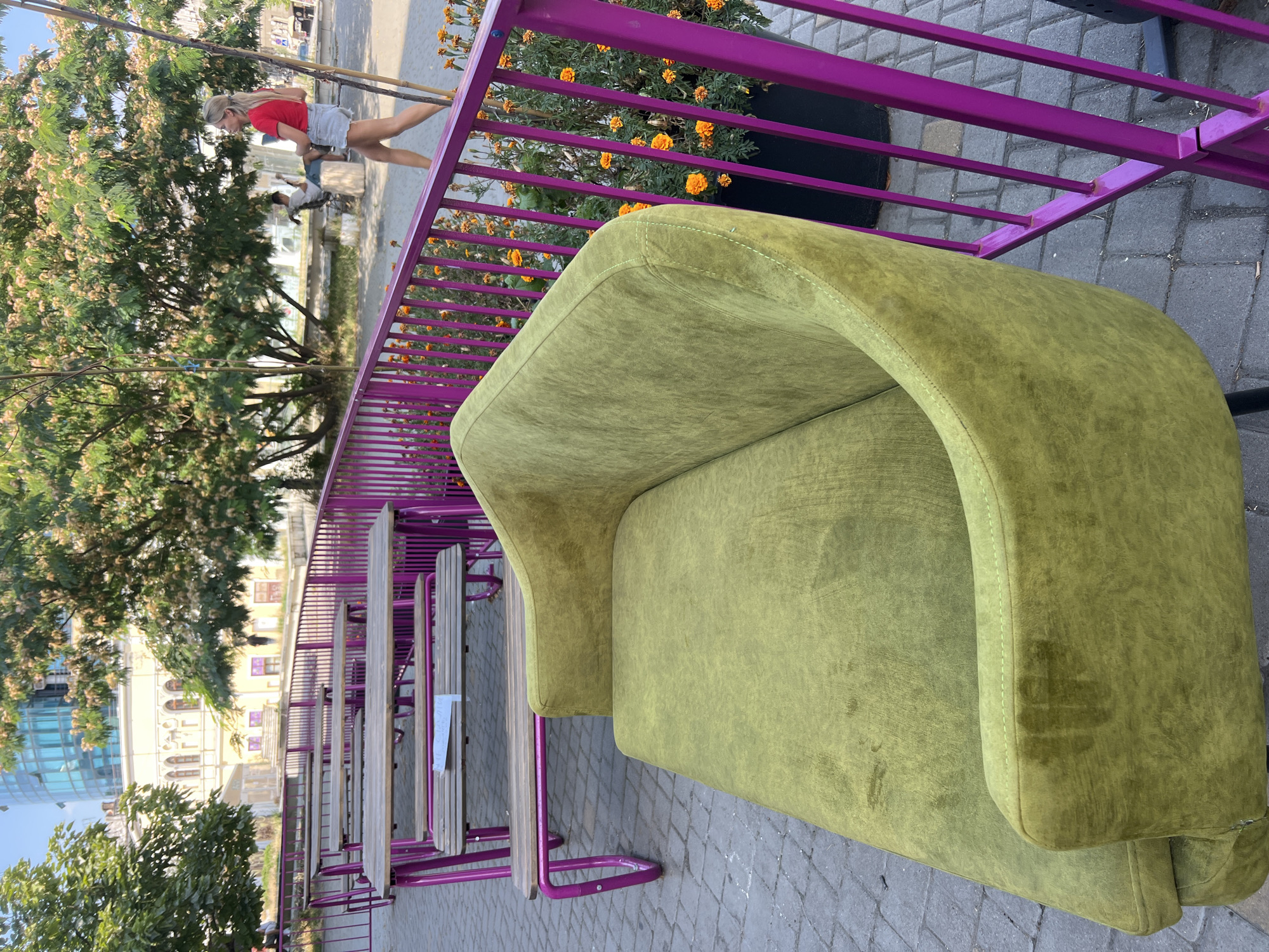 This is a street-level, outdoor shot of a cafe or restaurant seating area in Odesa, Ukraine, on a sunny day. The foreground is dominated by a plush, olive-green sofa. Behind the sofa, a bright magenta metal railing encloses the seating area, which is furnished with several wooden tables and benches with matching magenta metal frames. The tables are set up in a row, facing away from the viewer. A few trees with light-colored blossoms are visible in the background, casting dappled shadows. Behind the trees, buildings can be seen, including one with a curved, glass facade. A woman in a red top and denim shorts is walking away from the viewer, further down the street. The ground is paved with grey cobblestones. The scene depicts a pleasant, urban outdoor space.